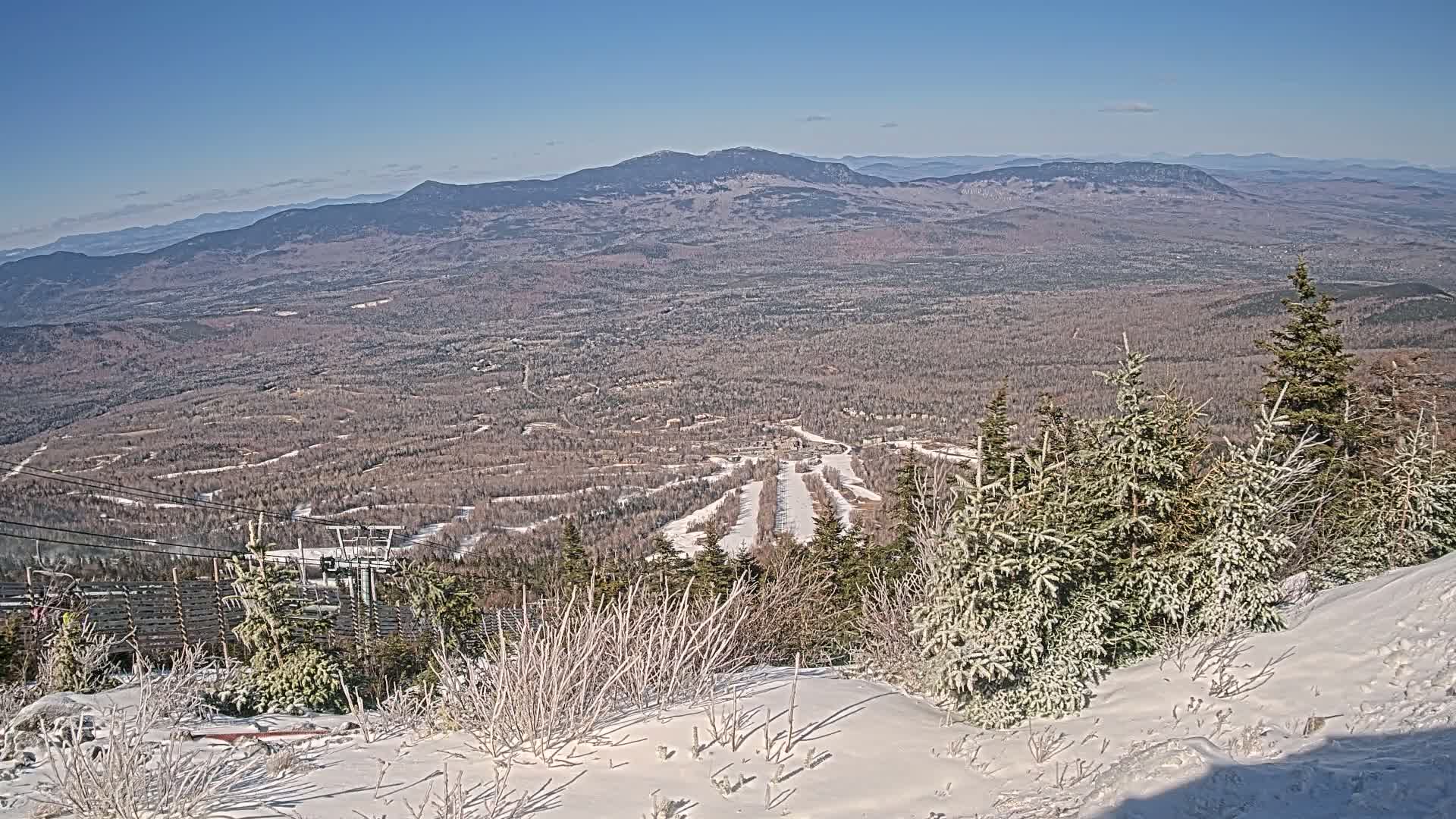 A snow-covered mountaintop overlooks a vast, snow-dusted forest and distant mountains under a clear blue sky.