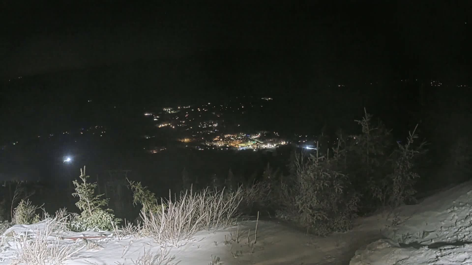 A snowy, nighttime view from a high vantage point shows a distant town illuminated by lights below.