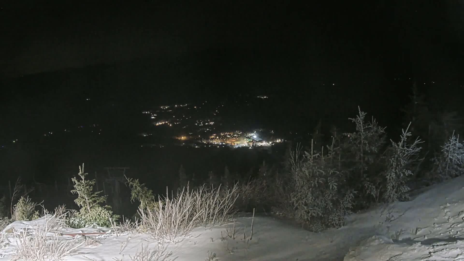 A snow-covered hillside at night overlooks a valley with scattered lights.