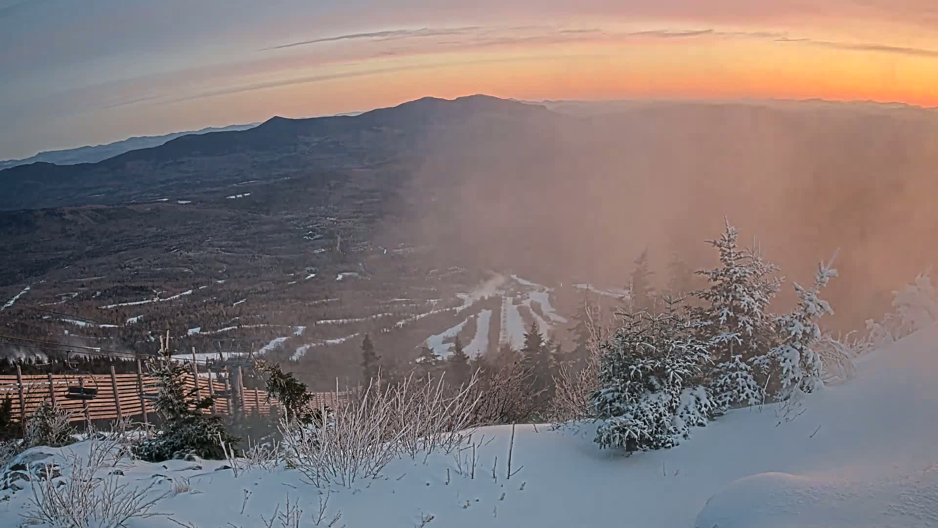 A snow-covered mountaintop overlooking a ski resort valley during a hazy, pinkish sunrise.
