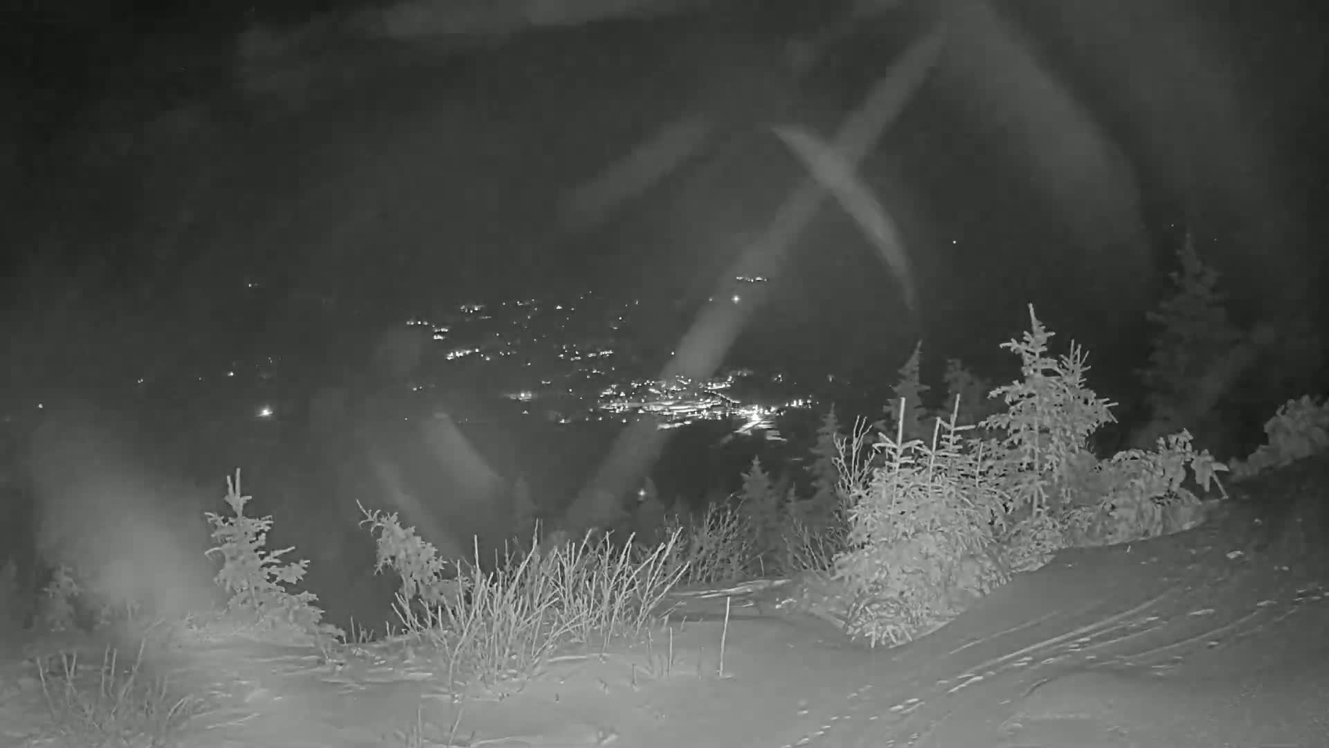 A nighttime, snowy scene shows a distant town's lights below a snow-covered hillside with evergreen trees, under a slightly hazy sky.