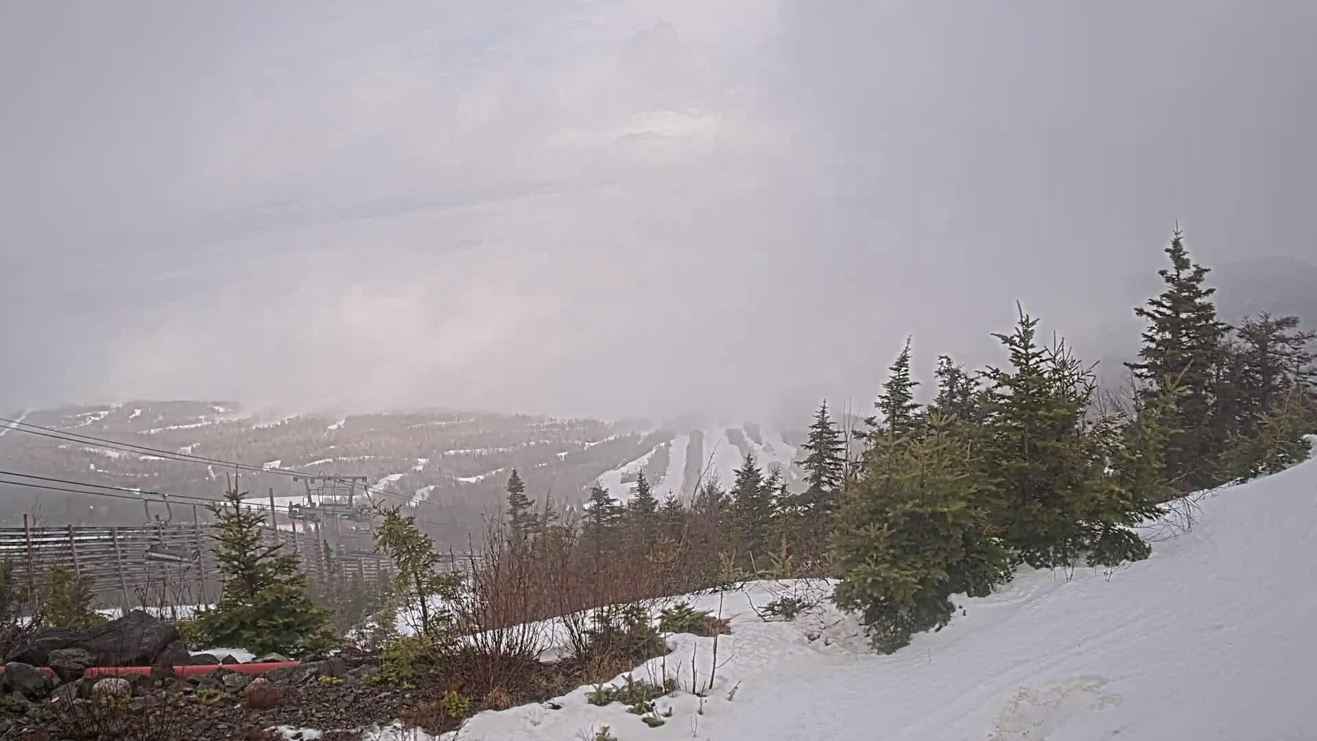A snow-covered ski resort is seen through a hazy, overcast sky, with evergreen trees in the foreground and a chairlift visible in the midground.