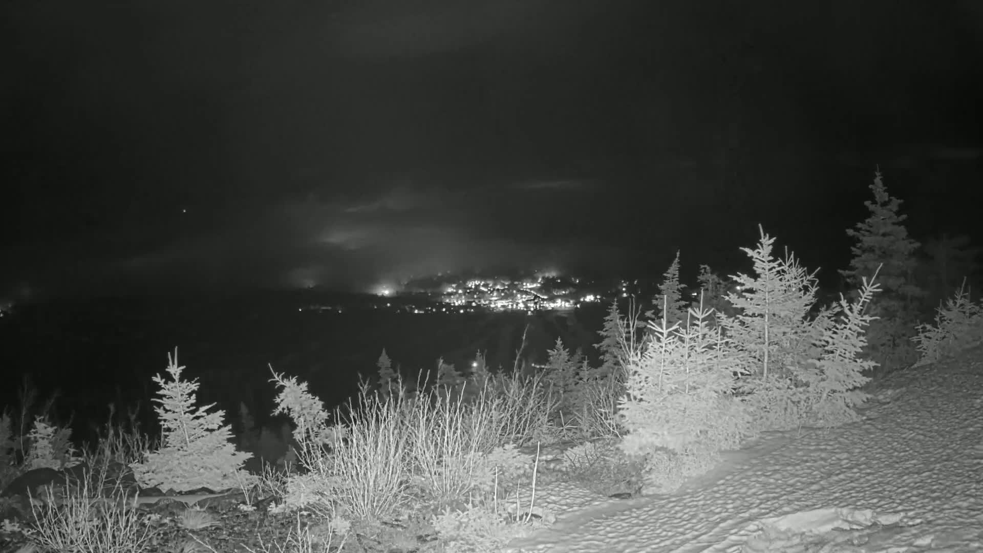 A nighttime view from a snow-covered hillside shows a valley with a town illuminated below, under a slightly cloudy sky.