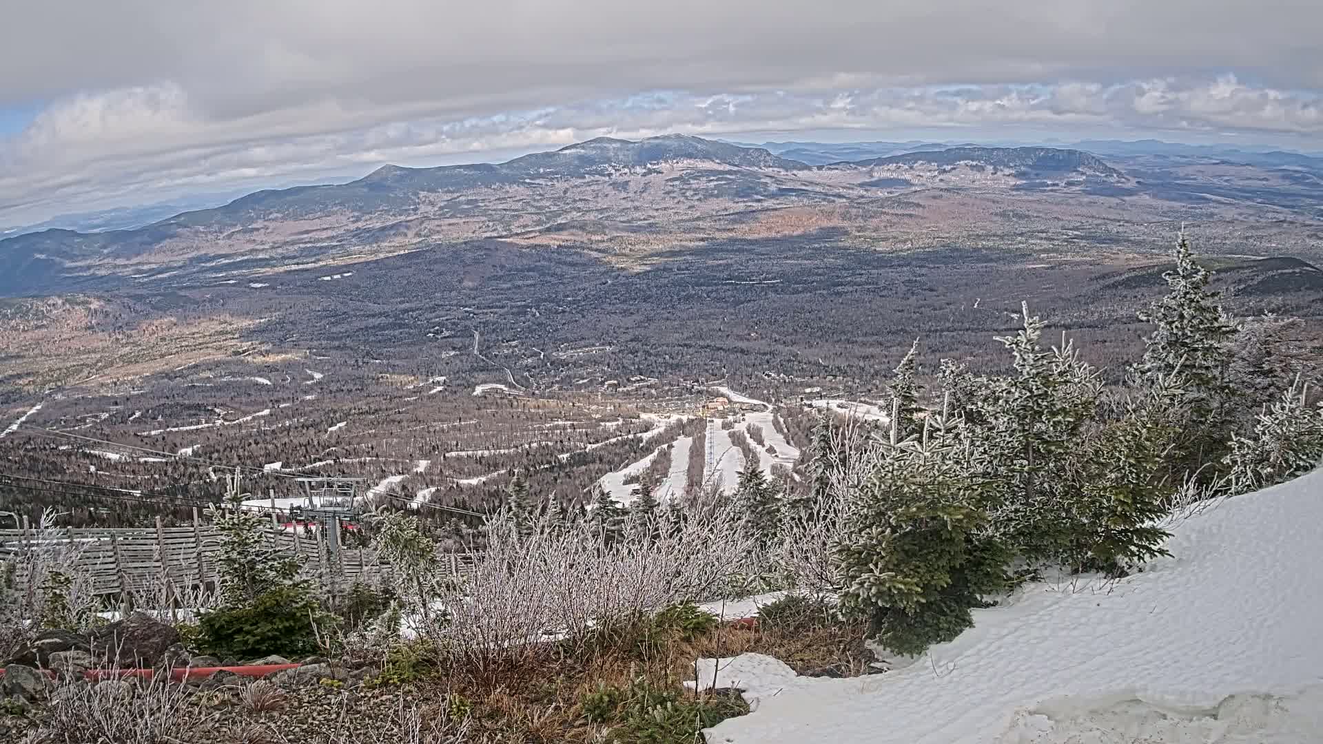 A snowy, mountainous landscape with a ski resort visible in the valley below under a cloudy sky.