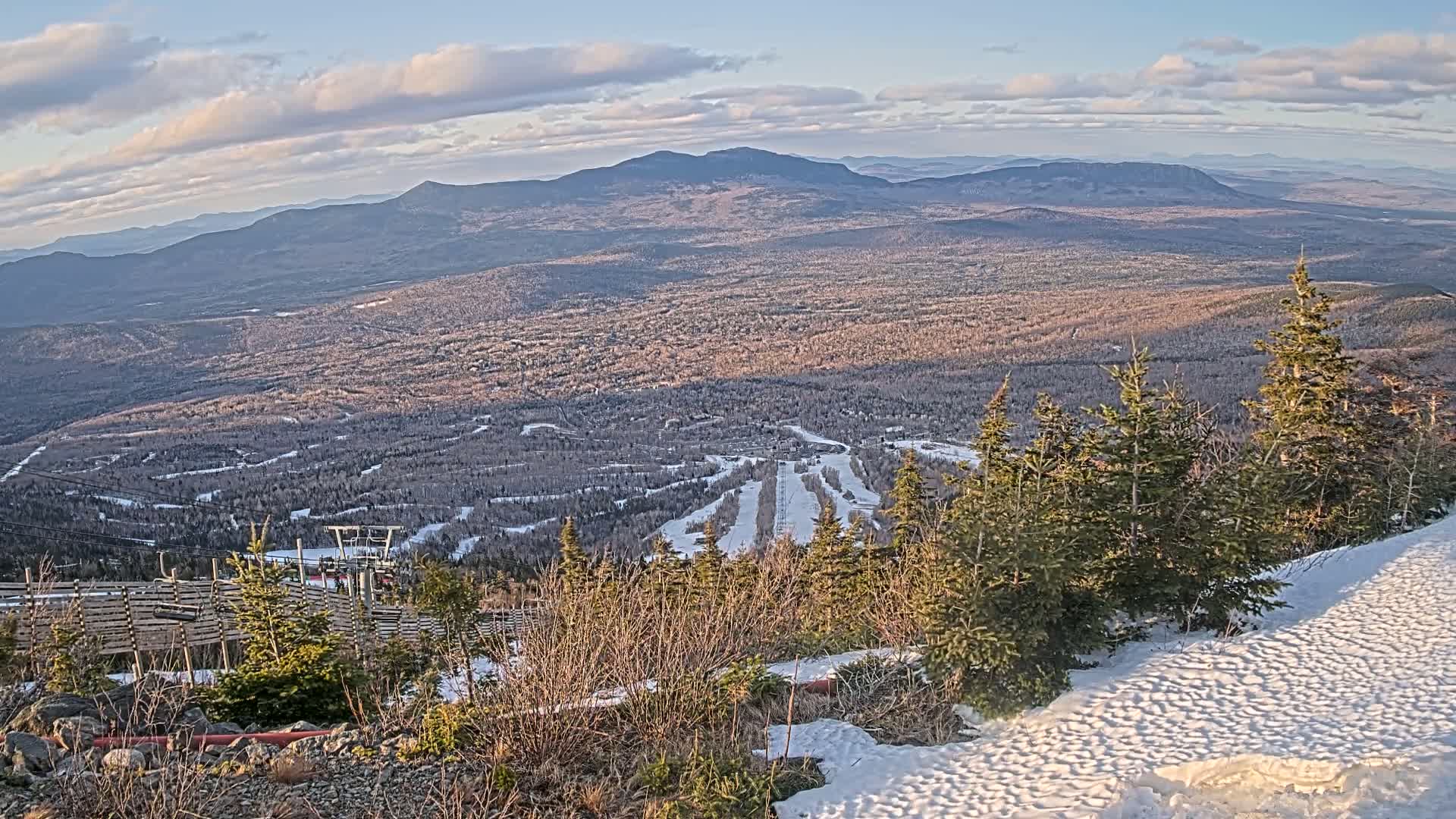 A snow-covered mountaintop overlooks a vast expanse of forested hills and valleys under a partly cloudy sky.