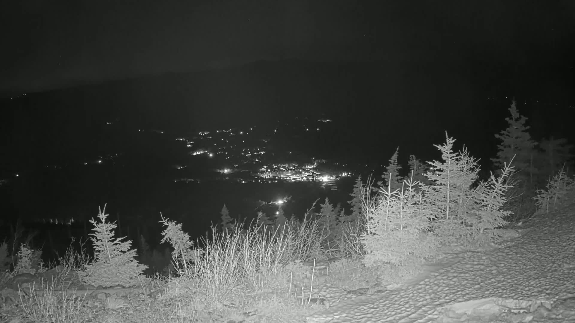 A nighttime view from a snow-covered hilltop shows a distant town illuminated by lights below.