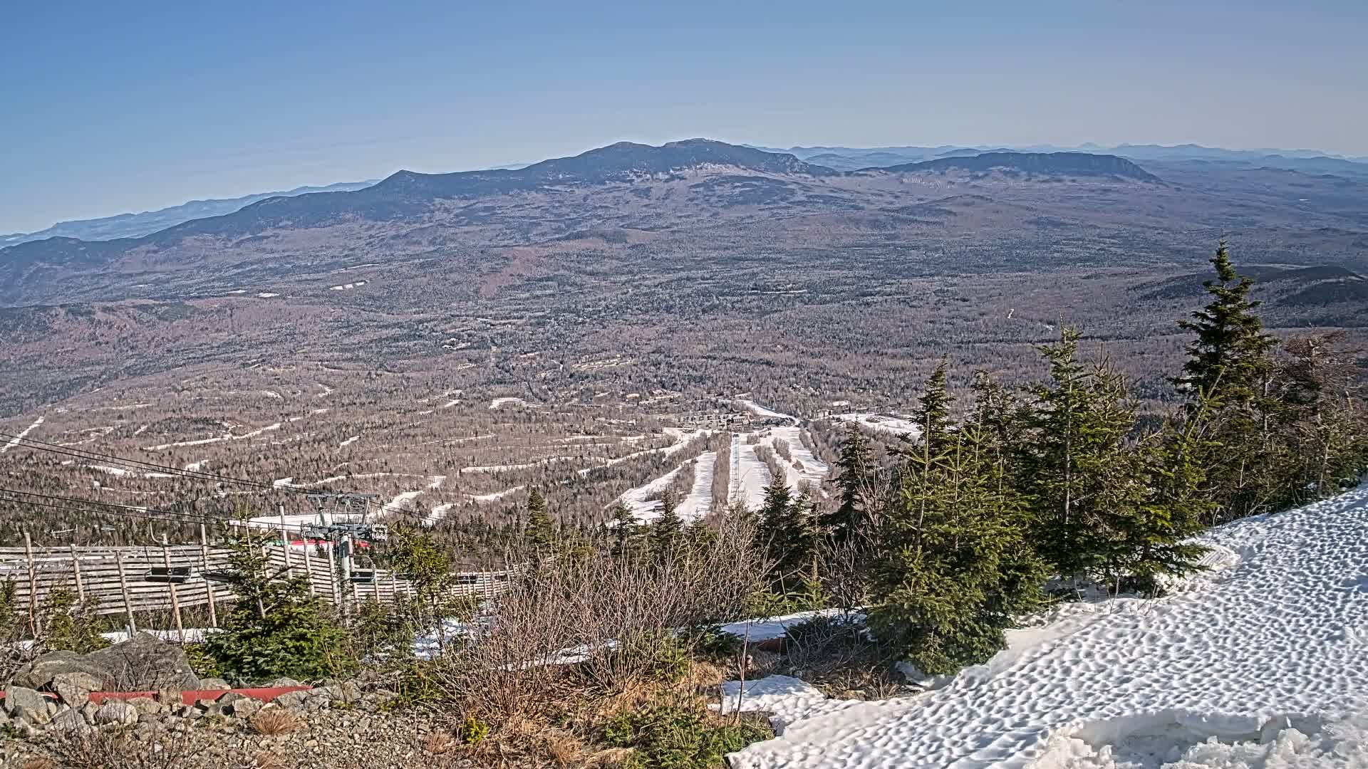 A snow-covered mountaintop provides a view of a vast valley with ski runs and distant mountains under a clear blue sky.