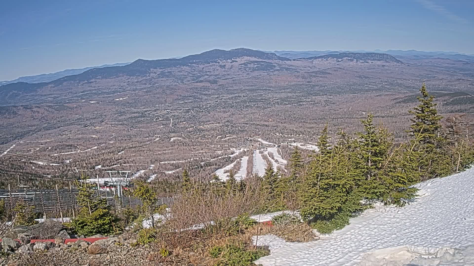 A snow-dusted mountaintop overlooks a vast expanse of forest and a ski resort under a clear blue sky.