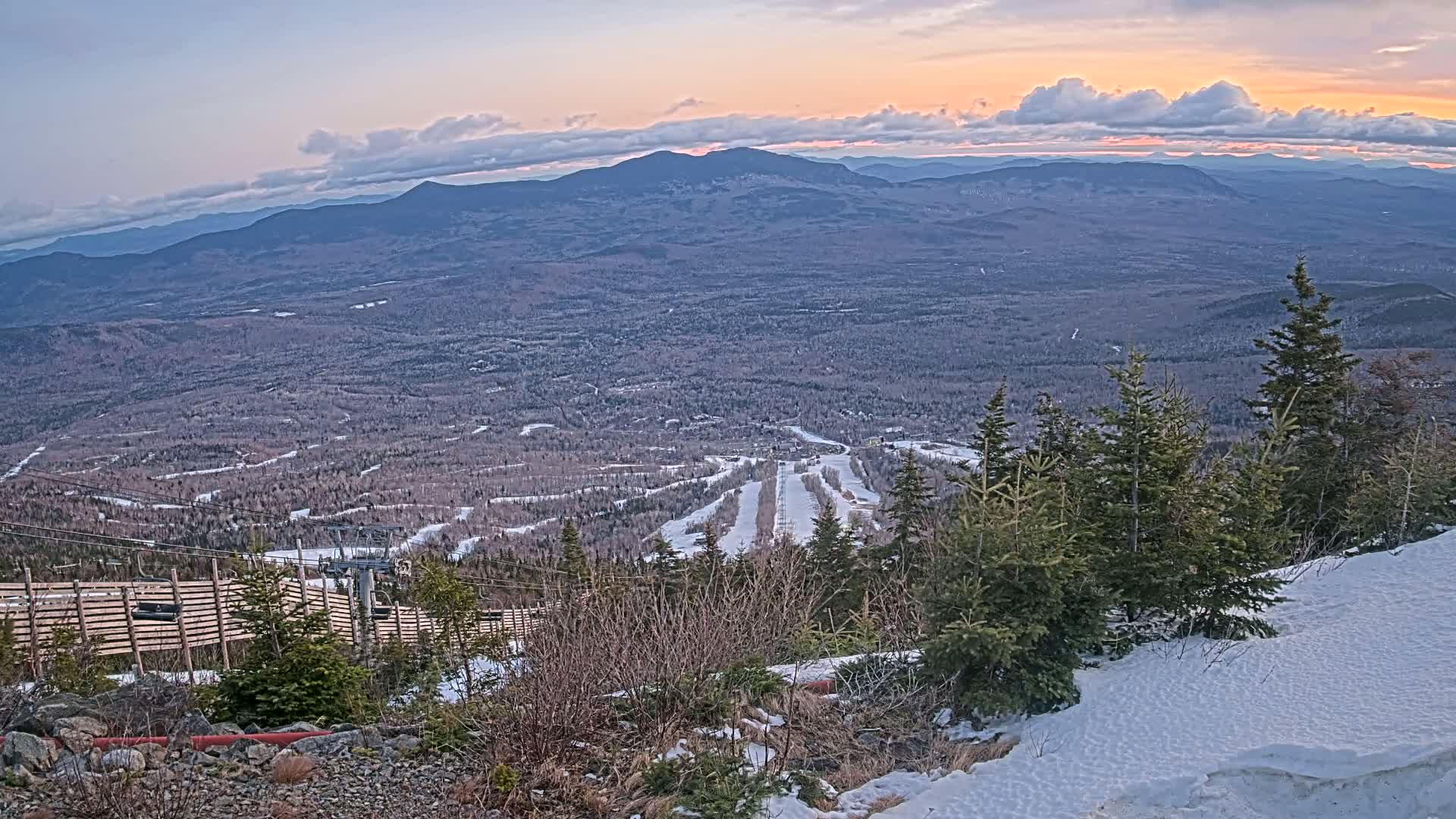 A high-angle view of a ski resort at sunset, showing snow-covered slopes, evergreen trees, and a vast mountain range in the background under a partly cloudy sky.