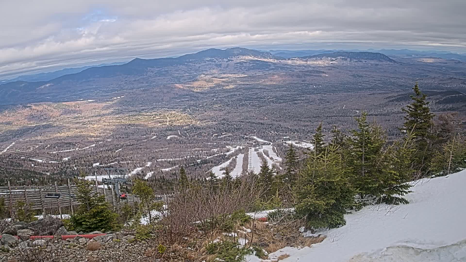 A high-angle, long shot shows a ski resort nestled in a valley, surrounded by snow-covered evergreens and distant, hazy mountains under a partly cloudy sky.