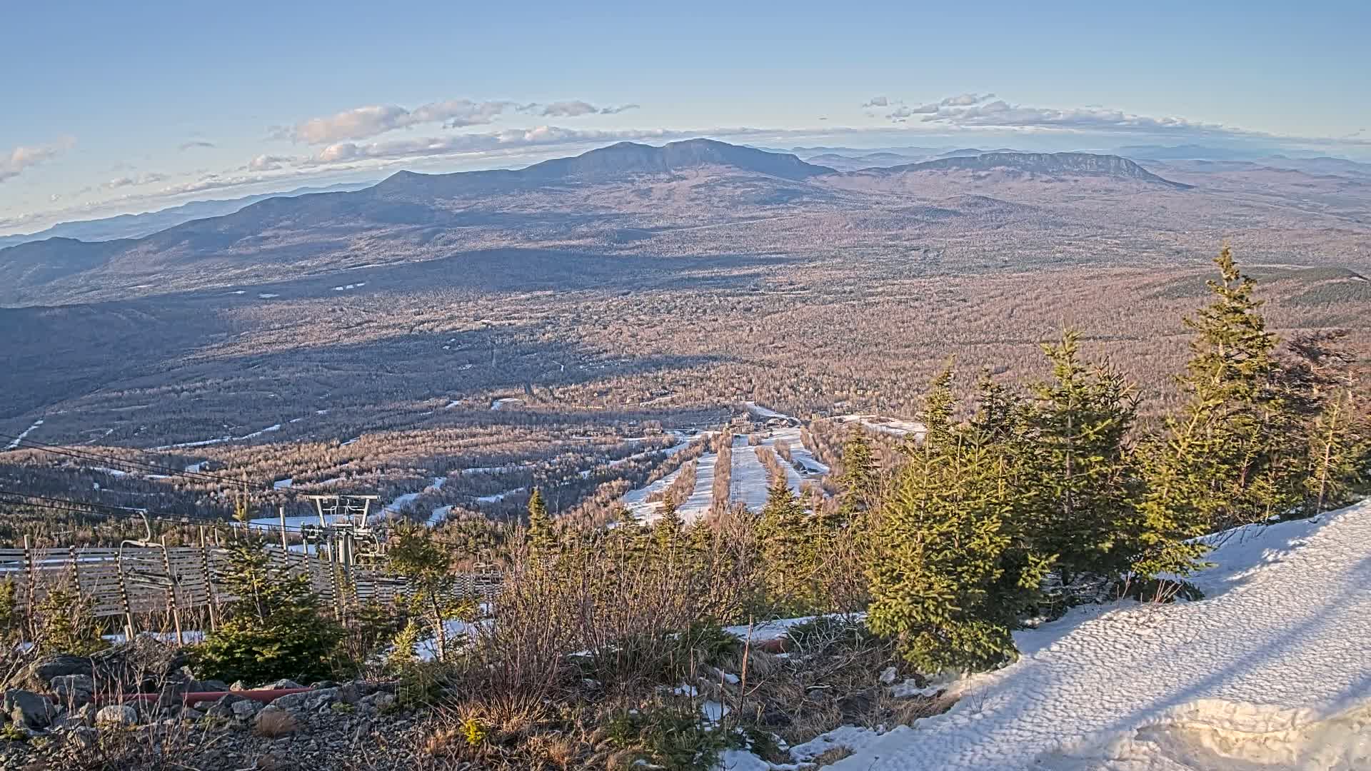A snowy mountaintop view overlooking a vast expanse of forest and distant mountains under a mostly clear, sunny sky.
