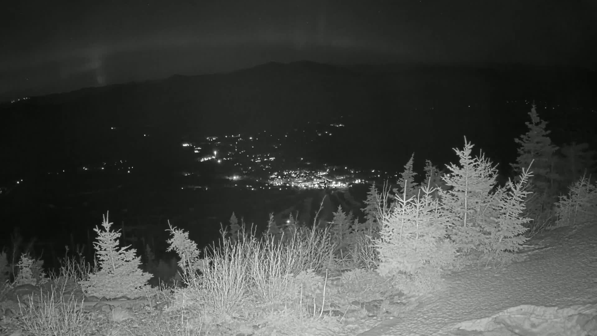 A nighttime view from a snowy mountaintop shows a valley town speckled with lights below, under a clear, dark sky.