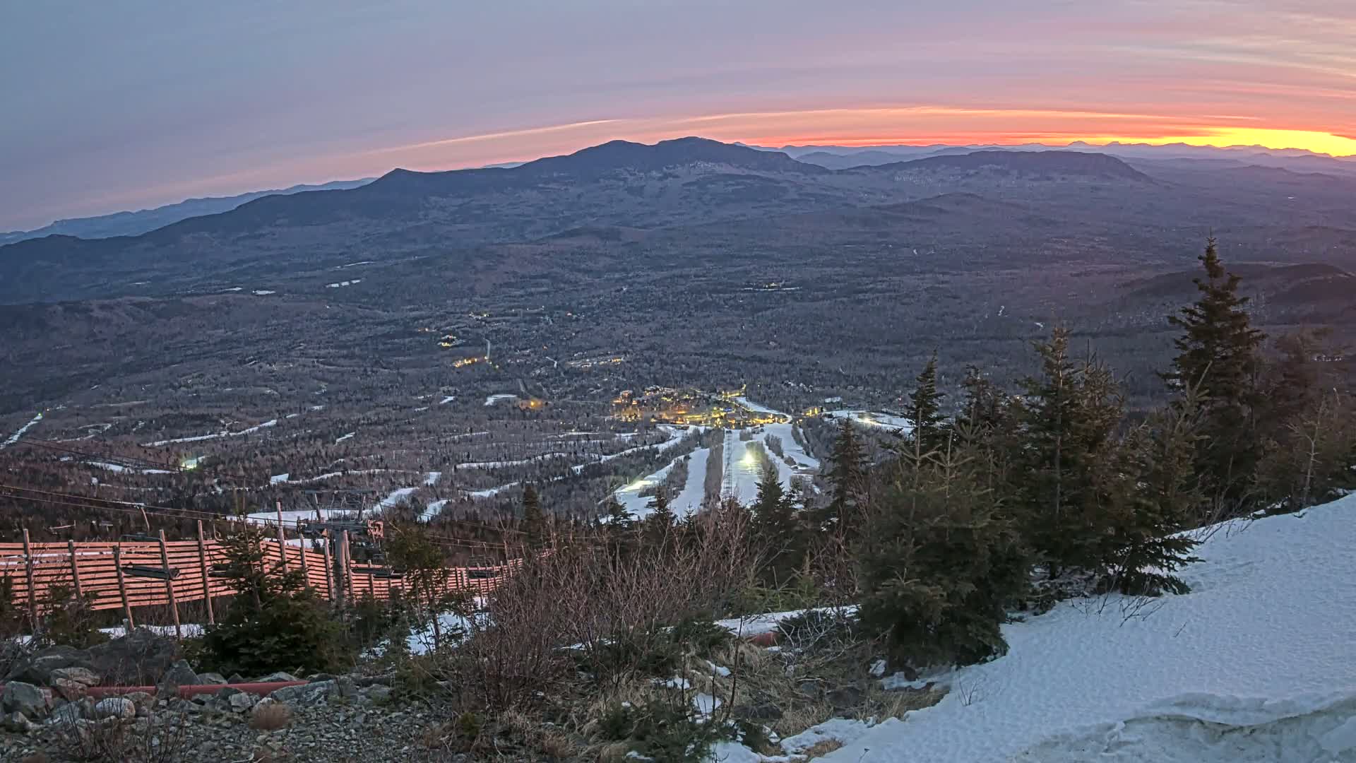 A snow-covered mountaintop at sunset overlooks a valley with a ski resort, illuminated by lights, under a twilight sky.