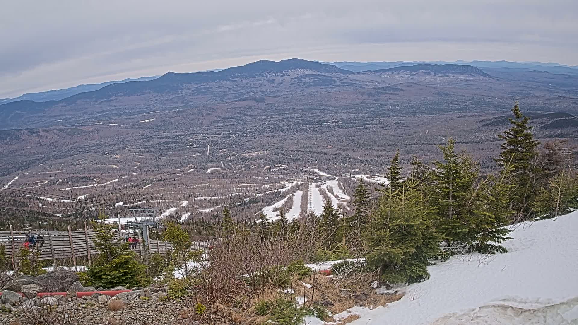 A snowy mountaintop ski resort overlooks a vast, snow-dusted forest and distant mountain range under an overcast sky.