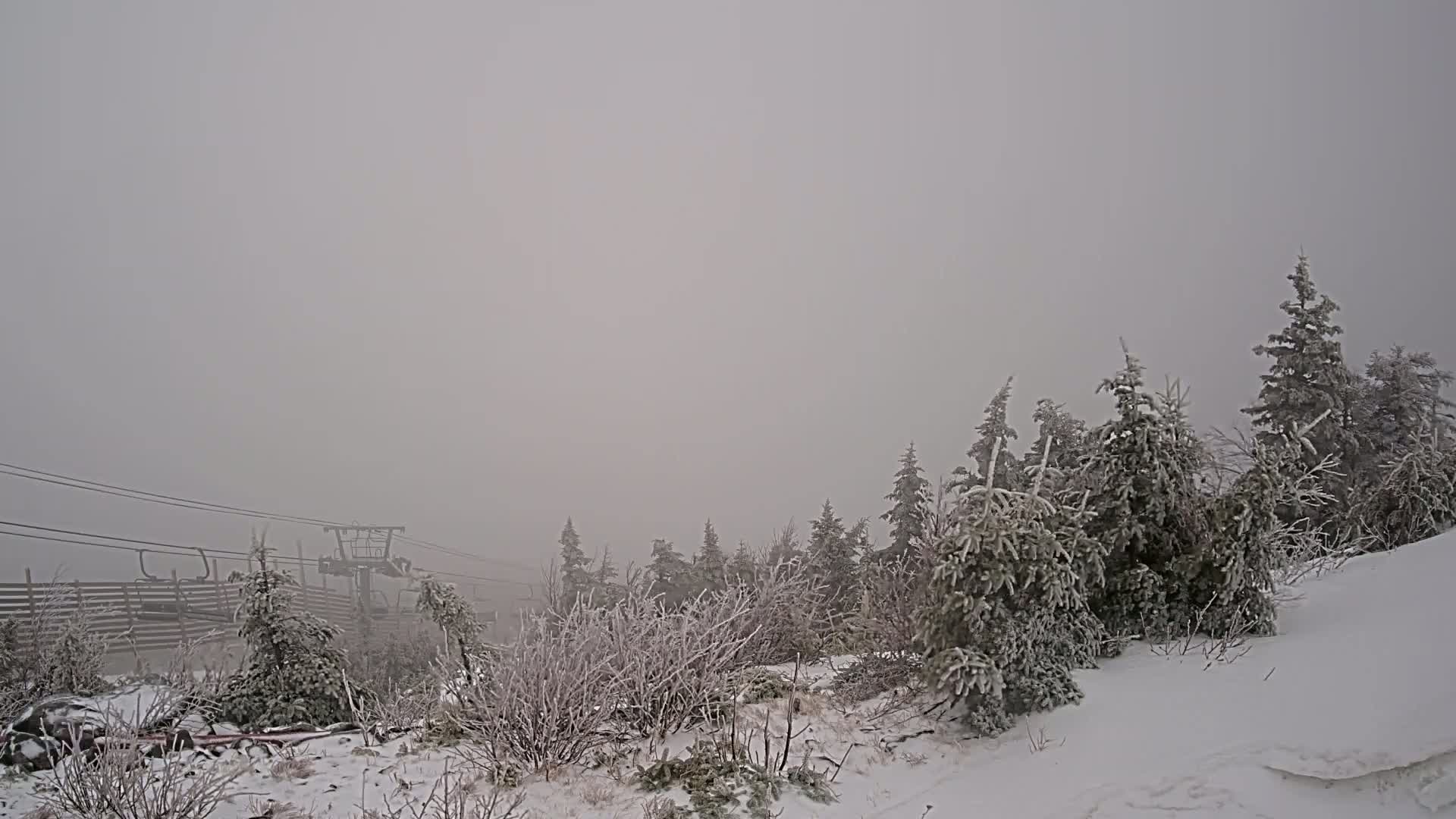 A snow-covered landscape with frost-covered trees and a chairlift in the distance is shrouded in heavy fog.