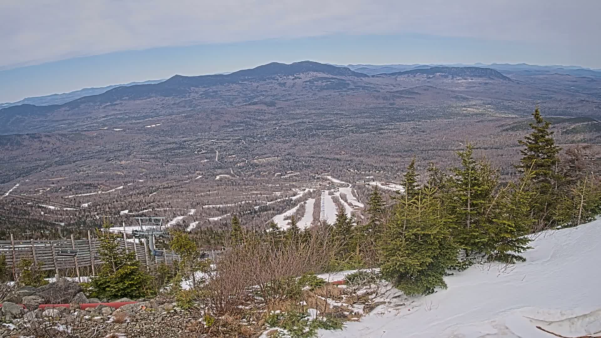 A snowy mountaintop view overlooking a vast expanse of forest with ski slopes and distant mountains under a cloudy sky.