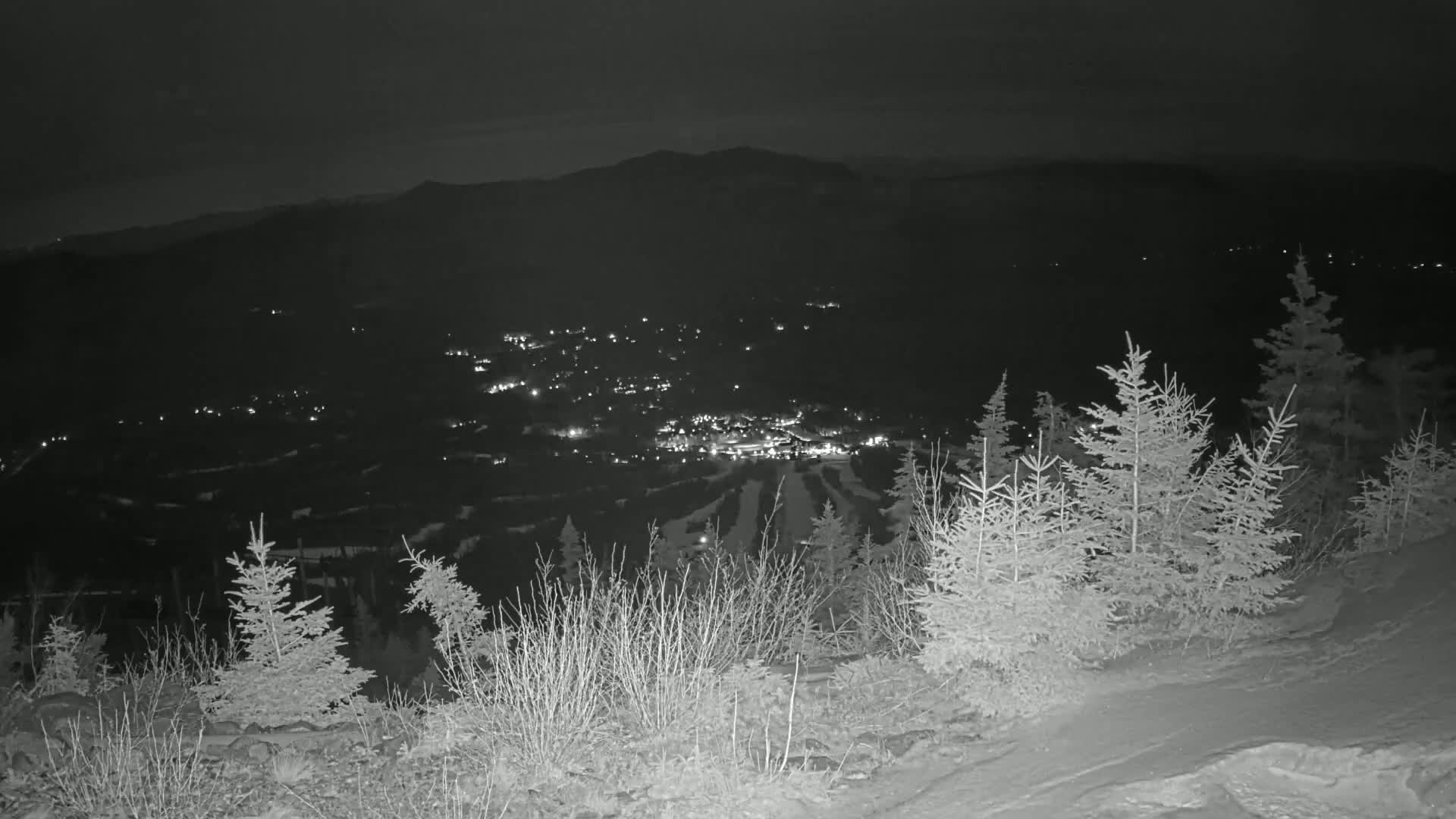 A nighttime, snowy mountaintop view overlooks a valley illuminated by scattered lights below.