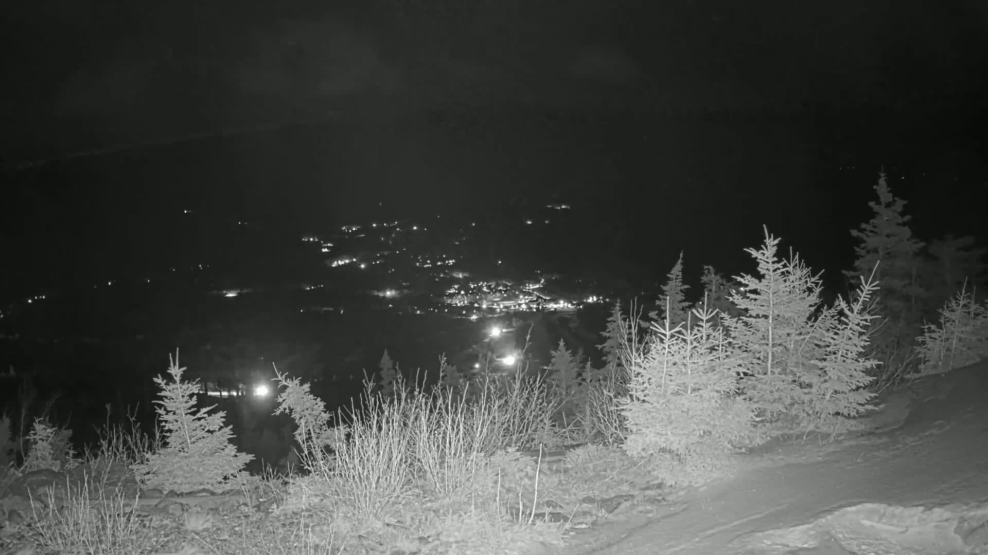 A nighttime, snow-covered hillside with evergreen trees overlooks a valley speckled with distant lights.