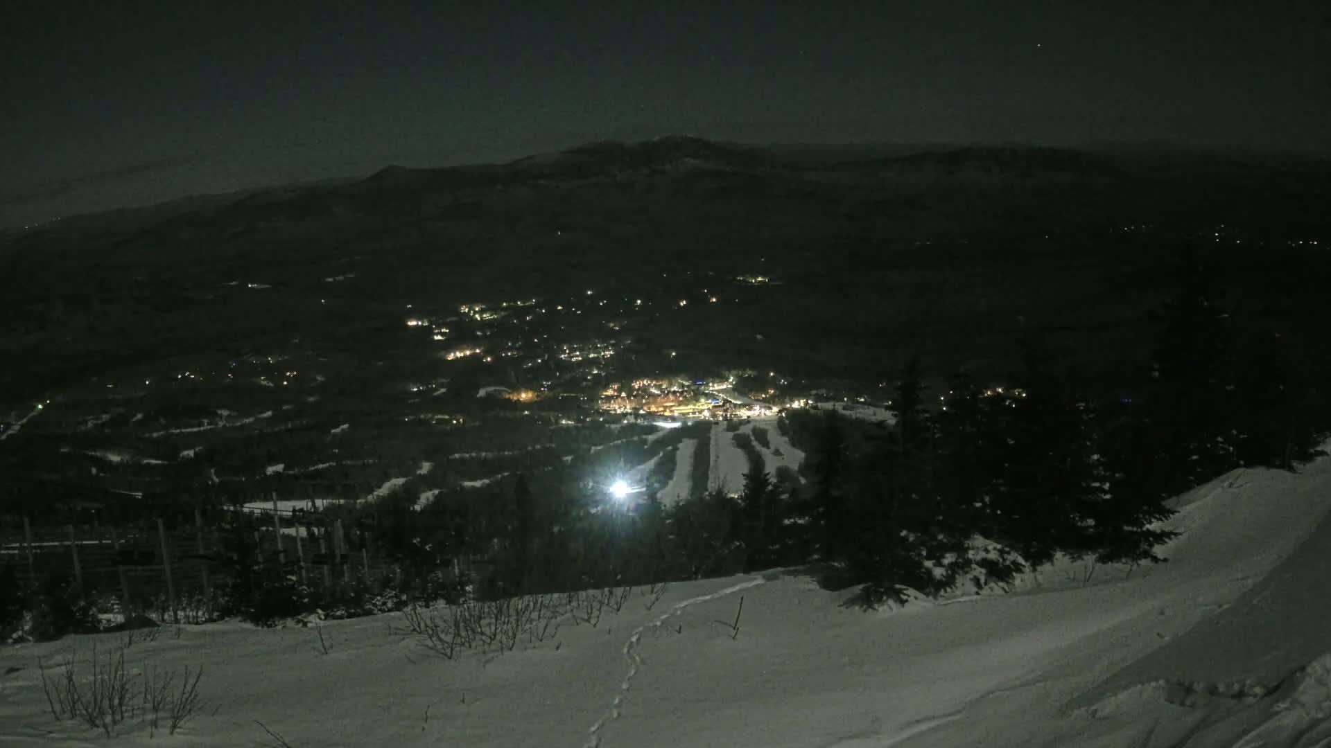 A snow-covered hillside at night overlooks a town and ski resort illuminated by many lights.