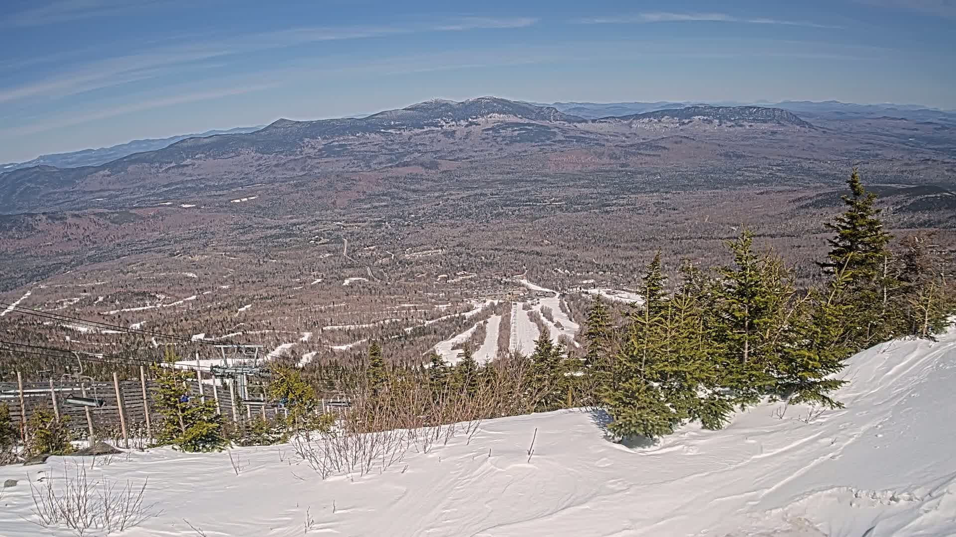 A snowy mountaintop view reveals a ski resort with runs and a chairlift, overlooking a vast expanse of forest and distant mountains under a clear blue sky.