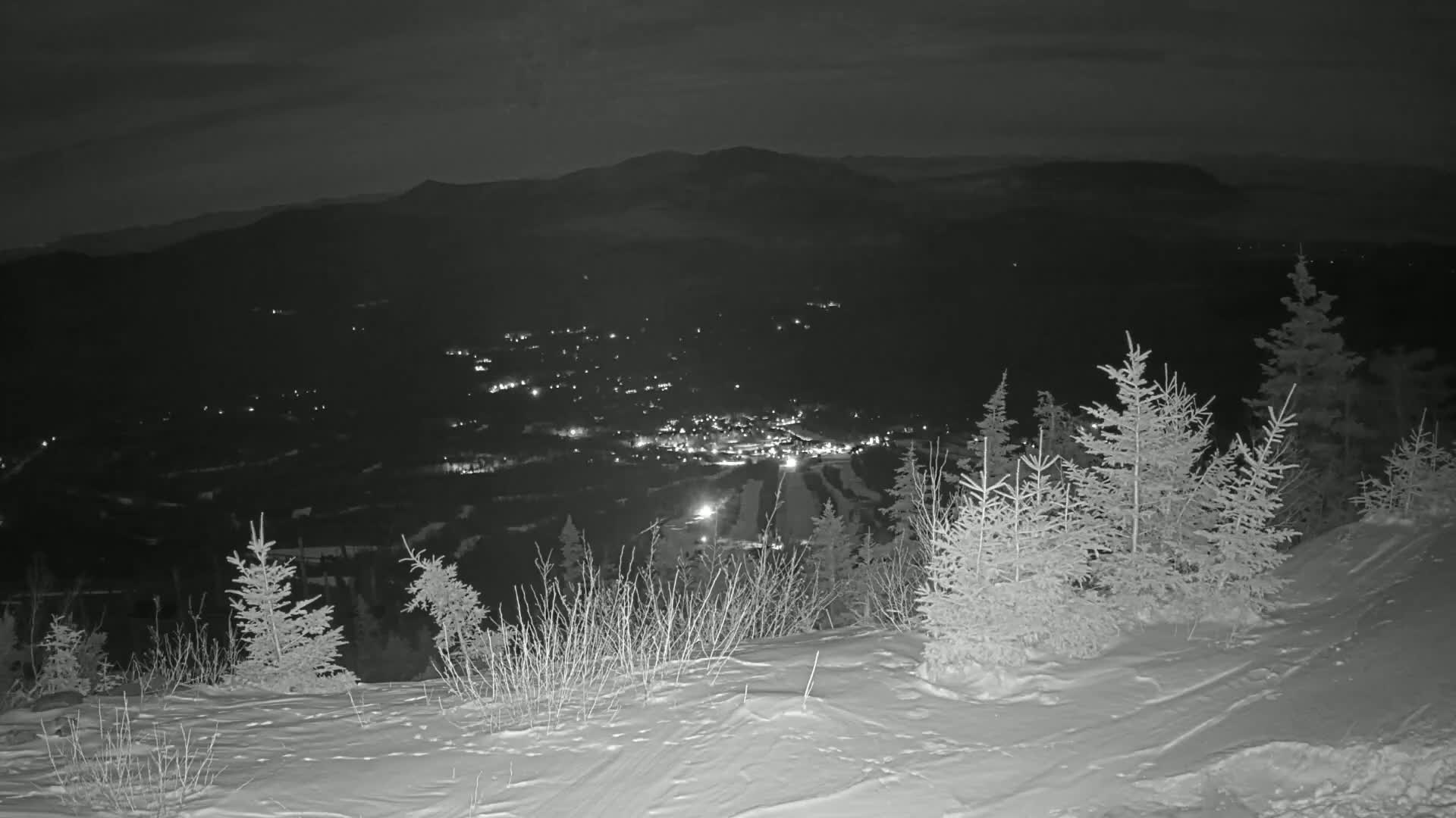 A snowy, nighttime mountaintop view overlooks a valley with a town illuminated below.