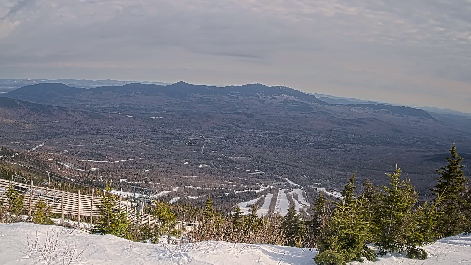 A snowy mountaintop overlooks a vast, wooded valley under a mostly cloudy sky.