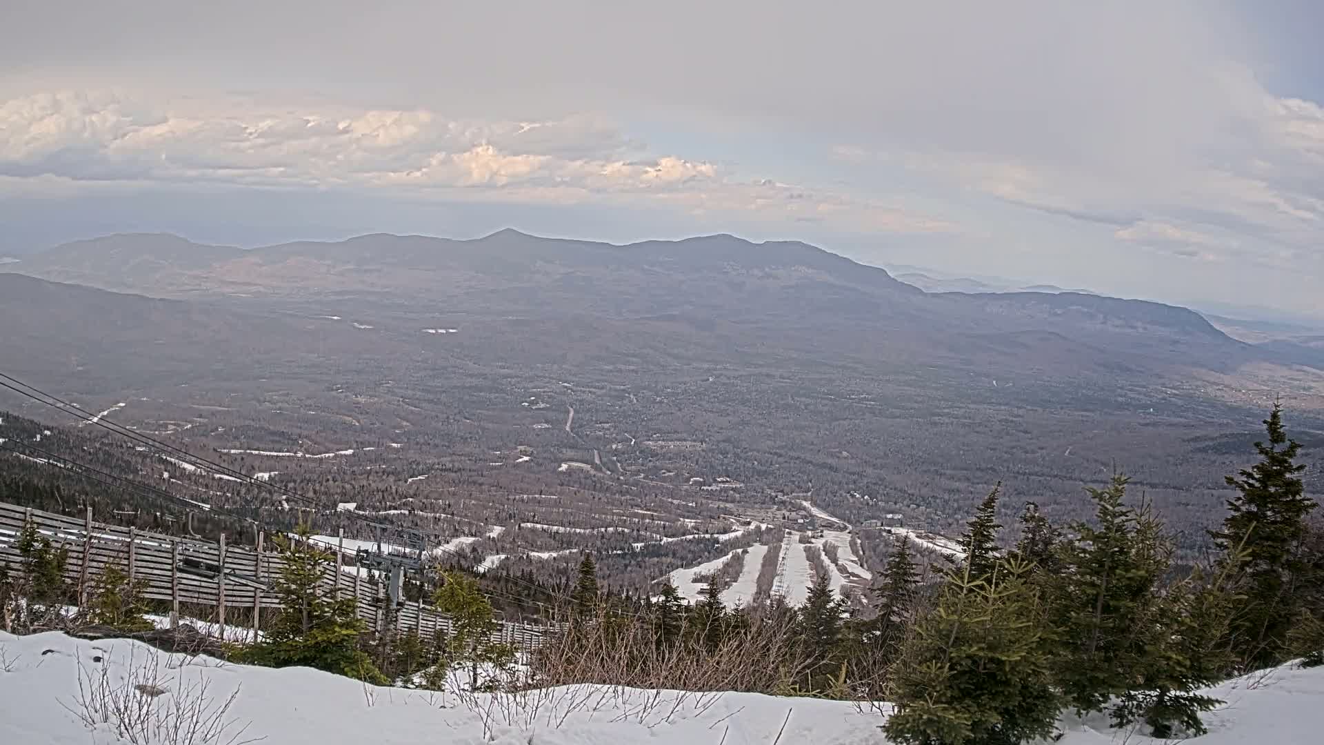 A snow-covered mountain slope overlooks a vast, tree-covered valley under a mostly cloudy sky.