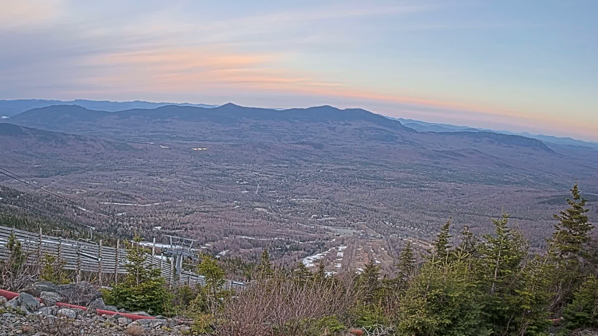 A mountaintop view at sunset reveals a vast valley of bare trees and a distant range of mountains under a pale sky.