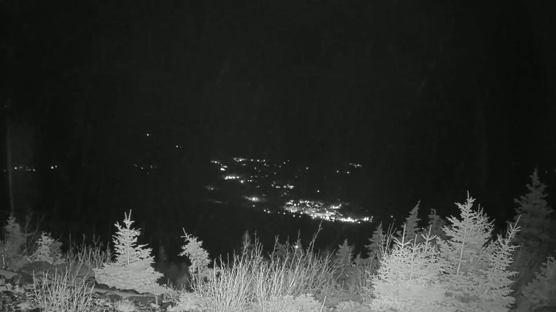 A nighttime view from a snow-covered hillside shows a distant town illuminated below, with light snowfall occurring.