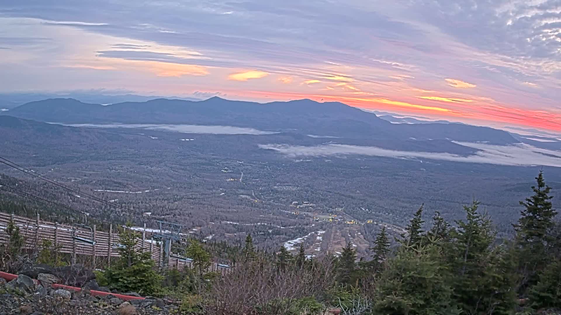 A mountaintop view at sunrise shows a valley covered in low clouds, with distant mountains silhouetted against a colorful sky.