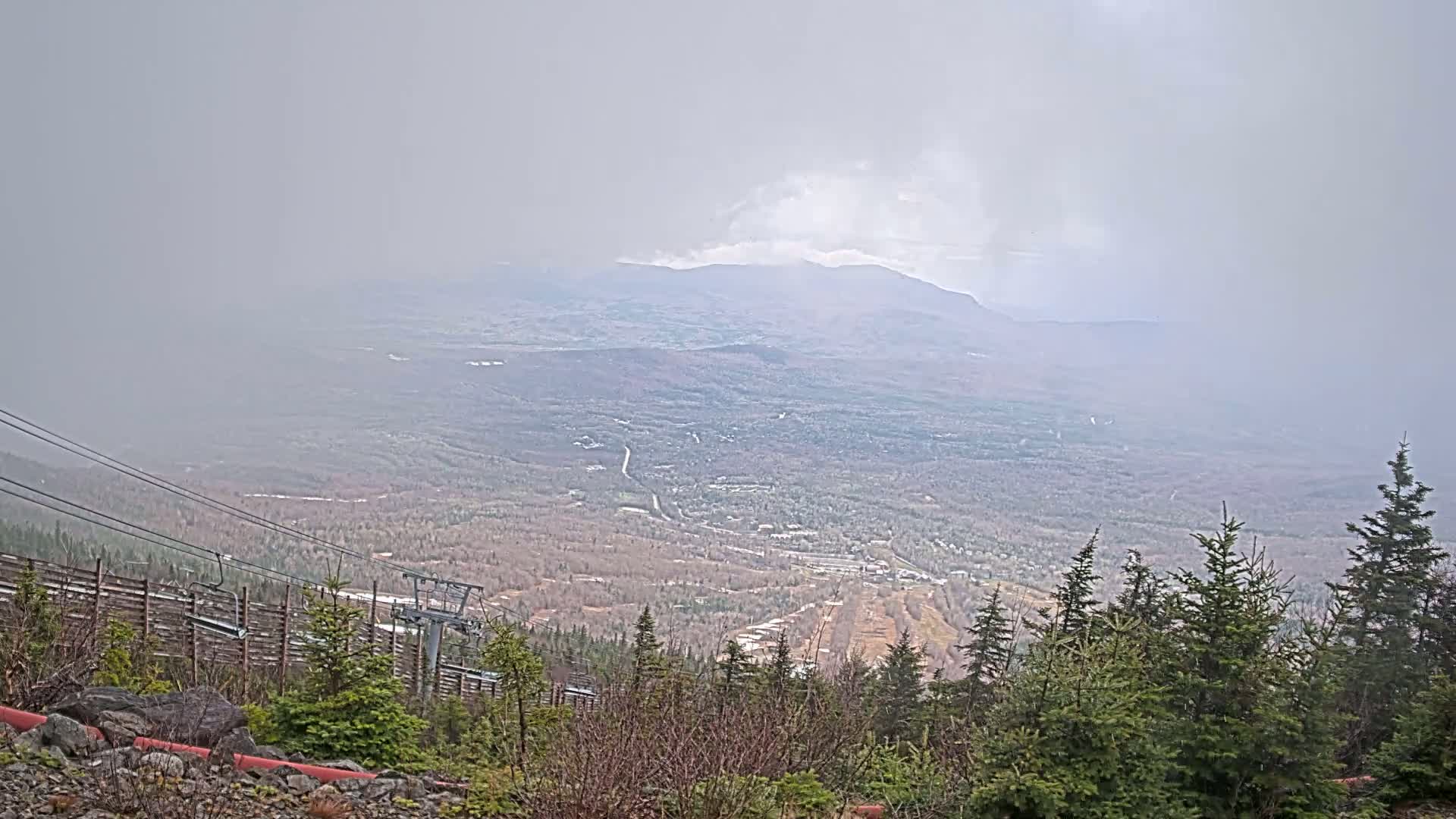 A hazy, overcast mountain vista reveals a valley with a town nestled among the trees and a ski lift in the foreground.