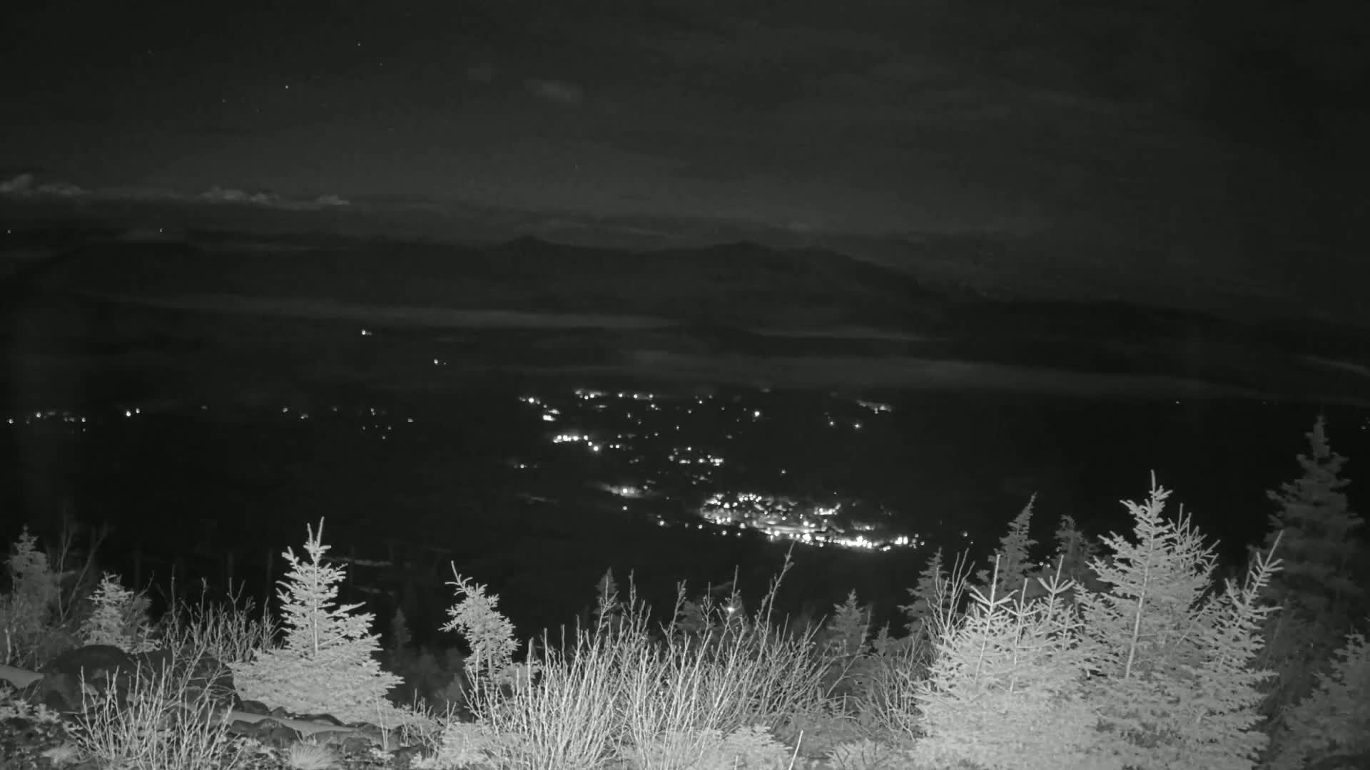 A nighttime view from a snow-covered mountaintop shows a valley below with a town illuminated by scattered lights, under a mostly clear, dark sky.