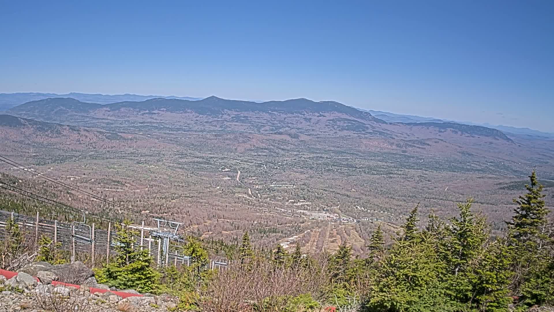 A vast, mountainous landscape stretches to the horizon under a clear blue sky, with a ski lift and evergreen trees in the foreground.