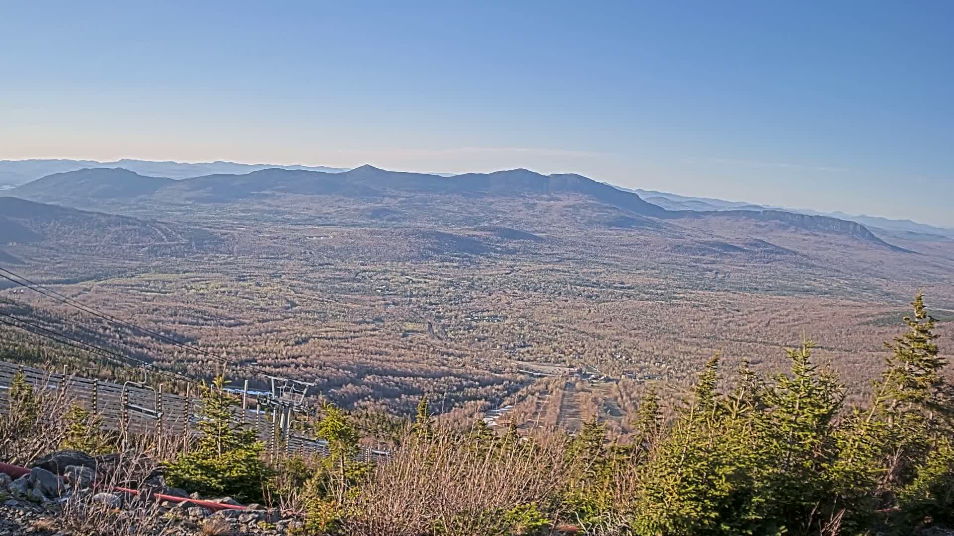 A vast expanse of tan-colored forest stretches out below a mountaintop viewpoint on a clear, sunny day, with distant blue mountains in the background and a ski lift partially visible in the foreground.