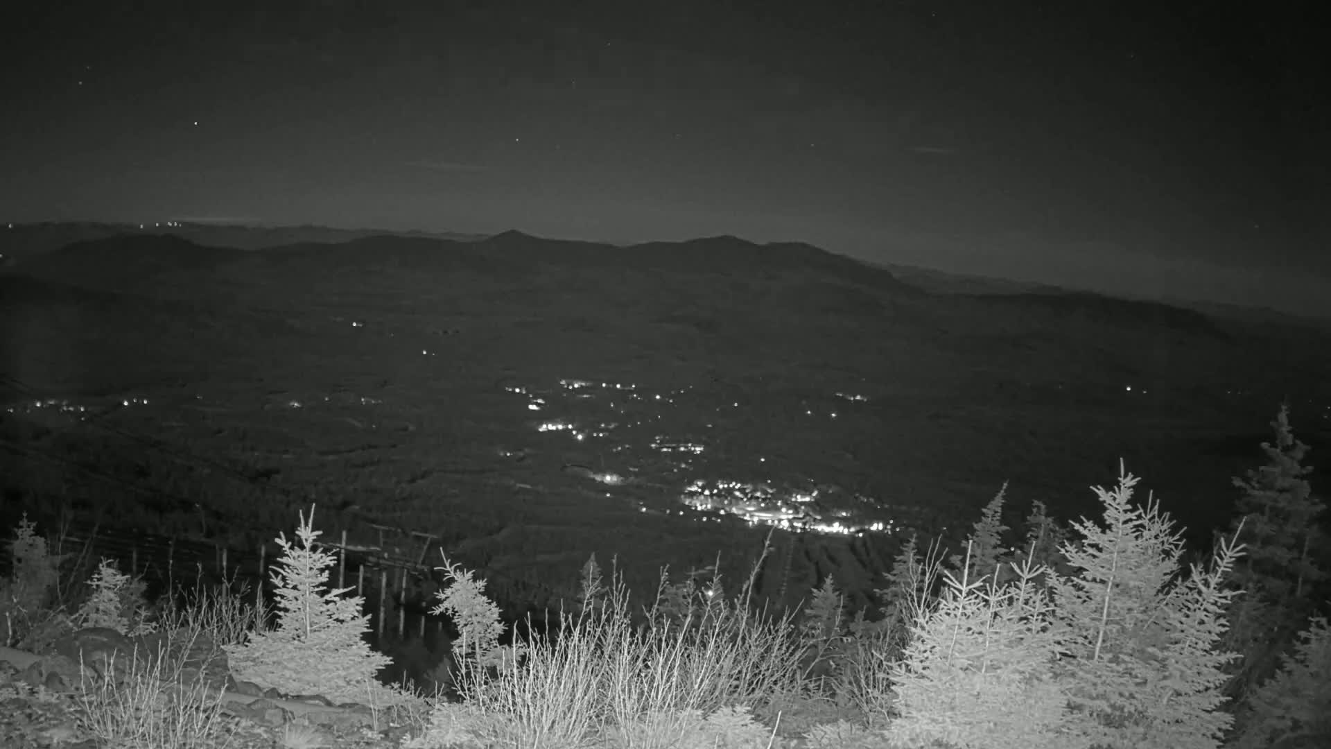A nighttime view from a mountaintop shows a valley with scattered lights below, and evergreen trees in the foreground under a clear, dark sky.