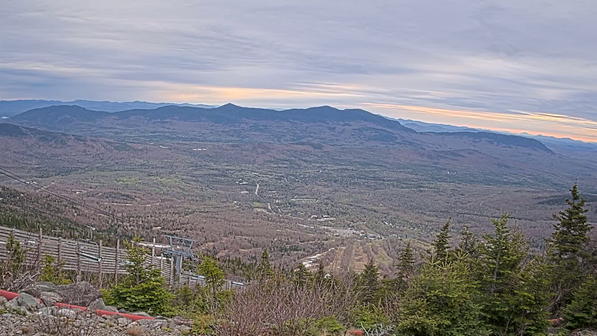 A panoramic view from a mountaintop shows a valley of forests and a small town under a partly cloudy, sunset sky.