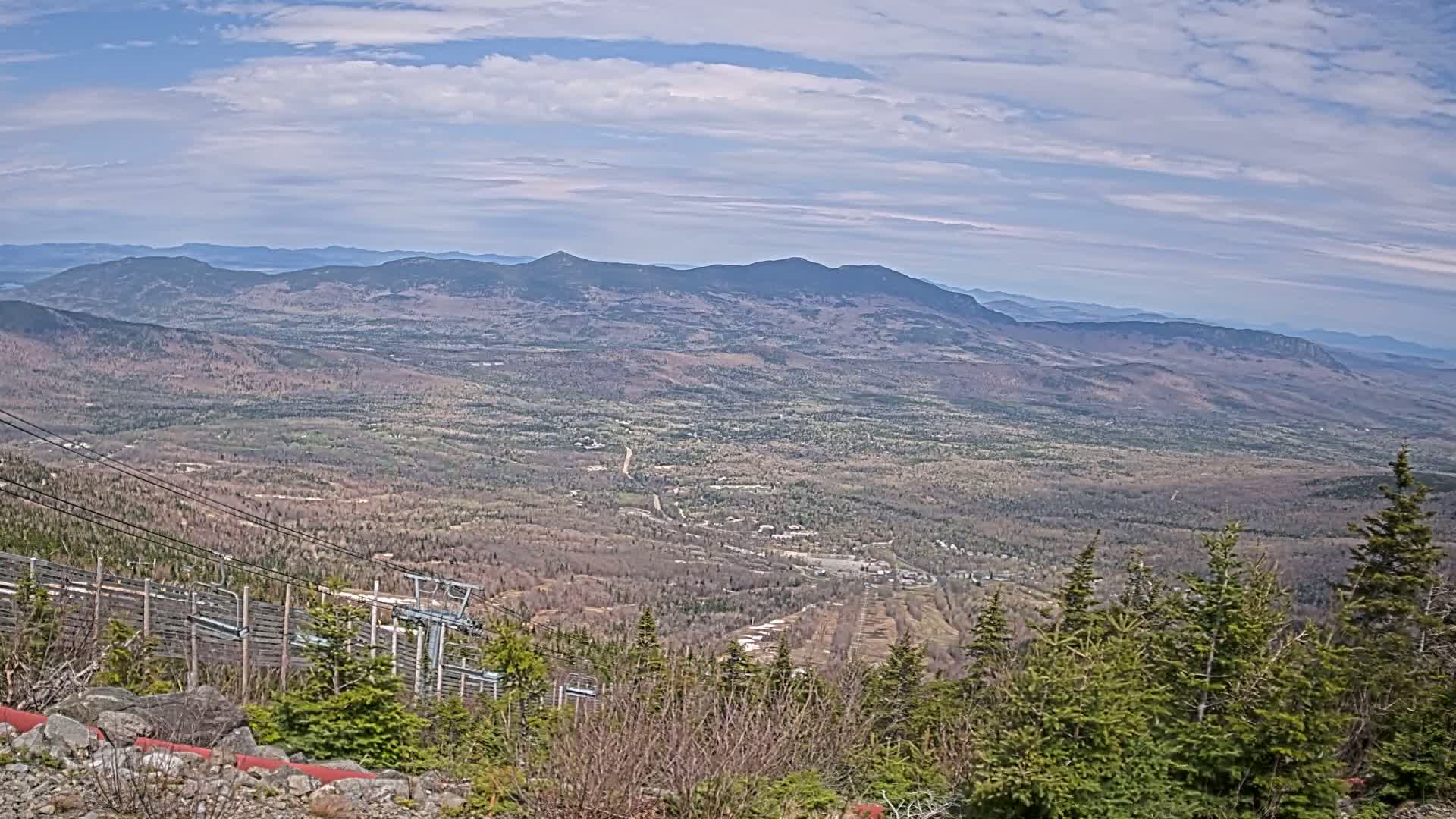 A vast, mountainous landscape stretches into the distance under a partly cloudy sky, with a ski lift and trees in the foreground.