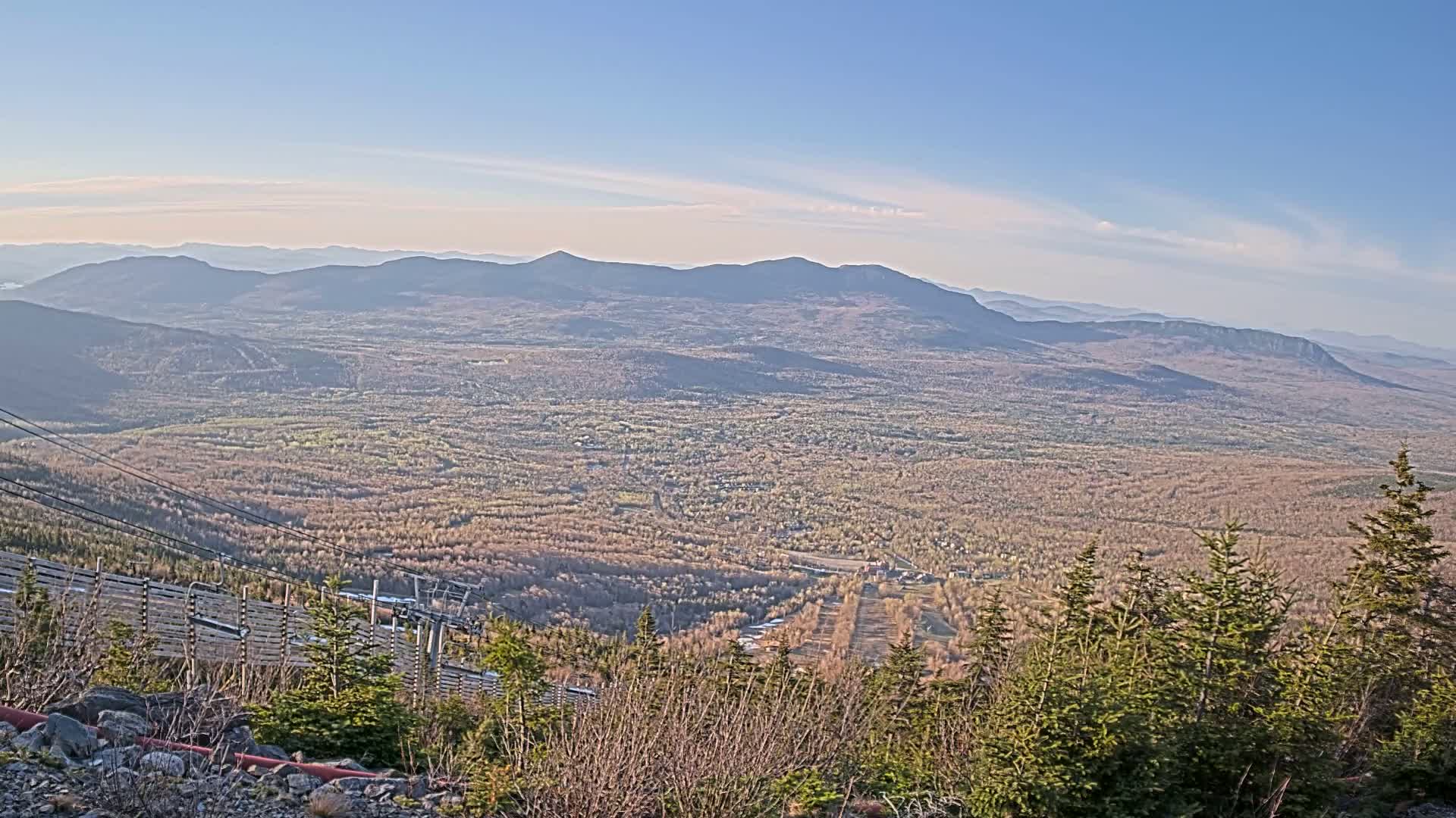 A vast, mountainous landscape stretches out under a clear, light-blue sky, featuring a valley covered in a mix of brown and green vegetation, with a ski lift and surrounding evergreens in the foreground.