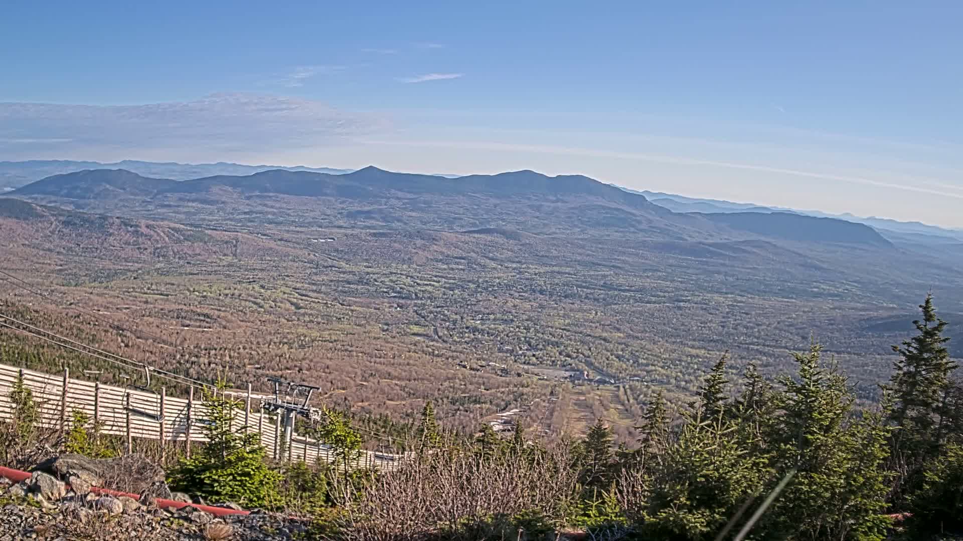 A vast, mountainous landscape stretches out under a clear blue sky, revealing a valley of trees and a ski lift in the foreground.