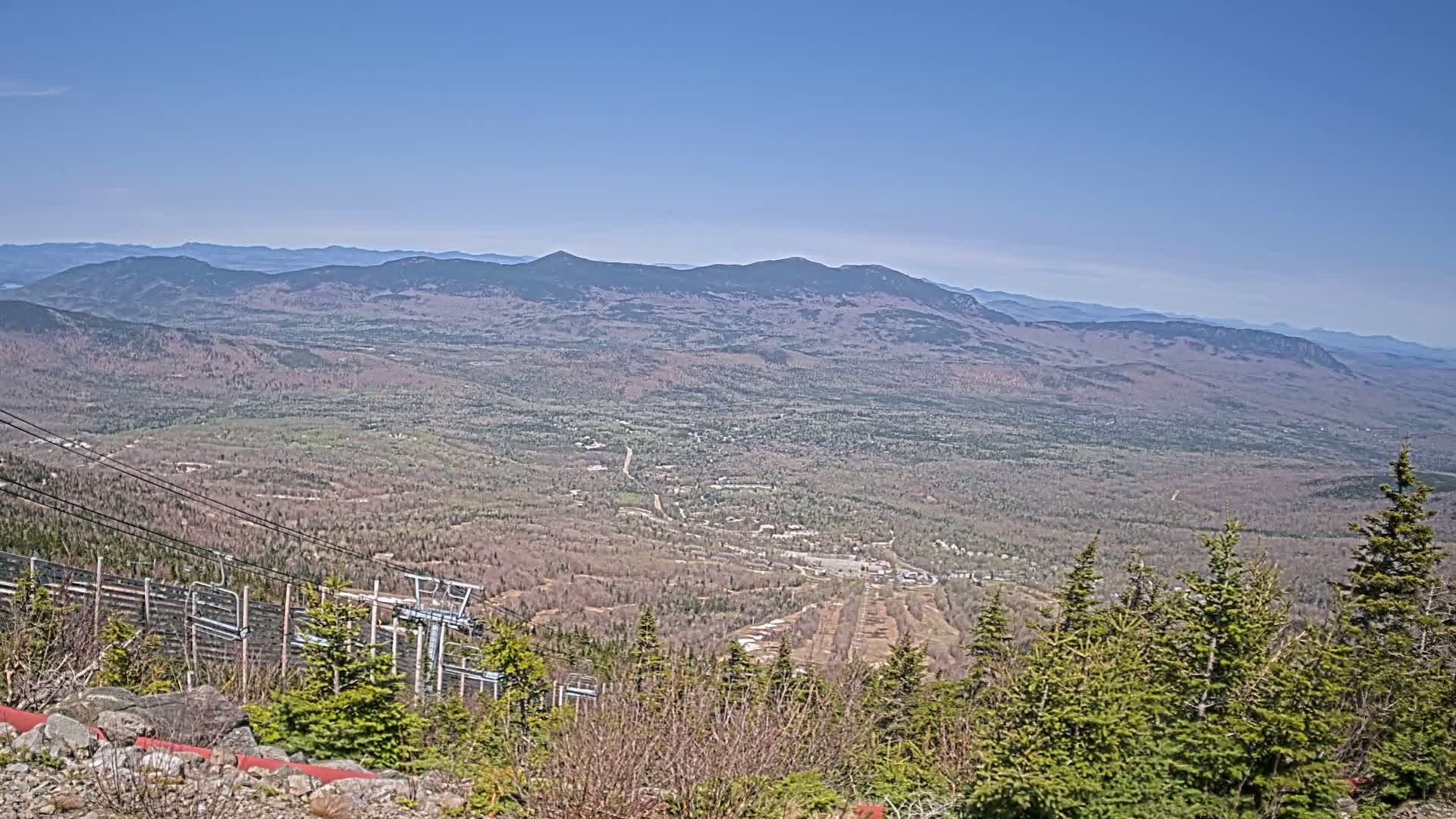 A vast, mountainous landscape stretches out under a clear blue sky, showing a mix of evergreens and deciduous trees in shades of brown and green, with a ski lift visible in the foreground.