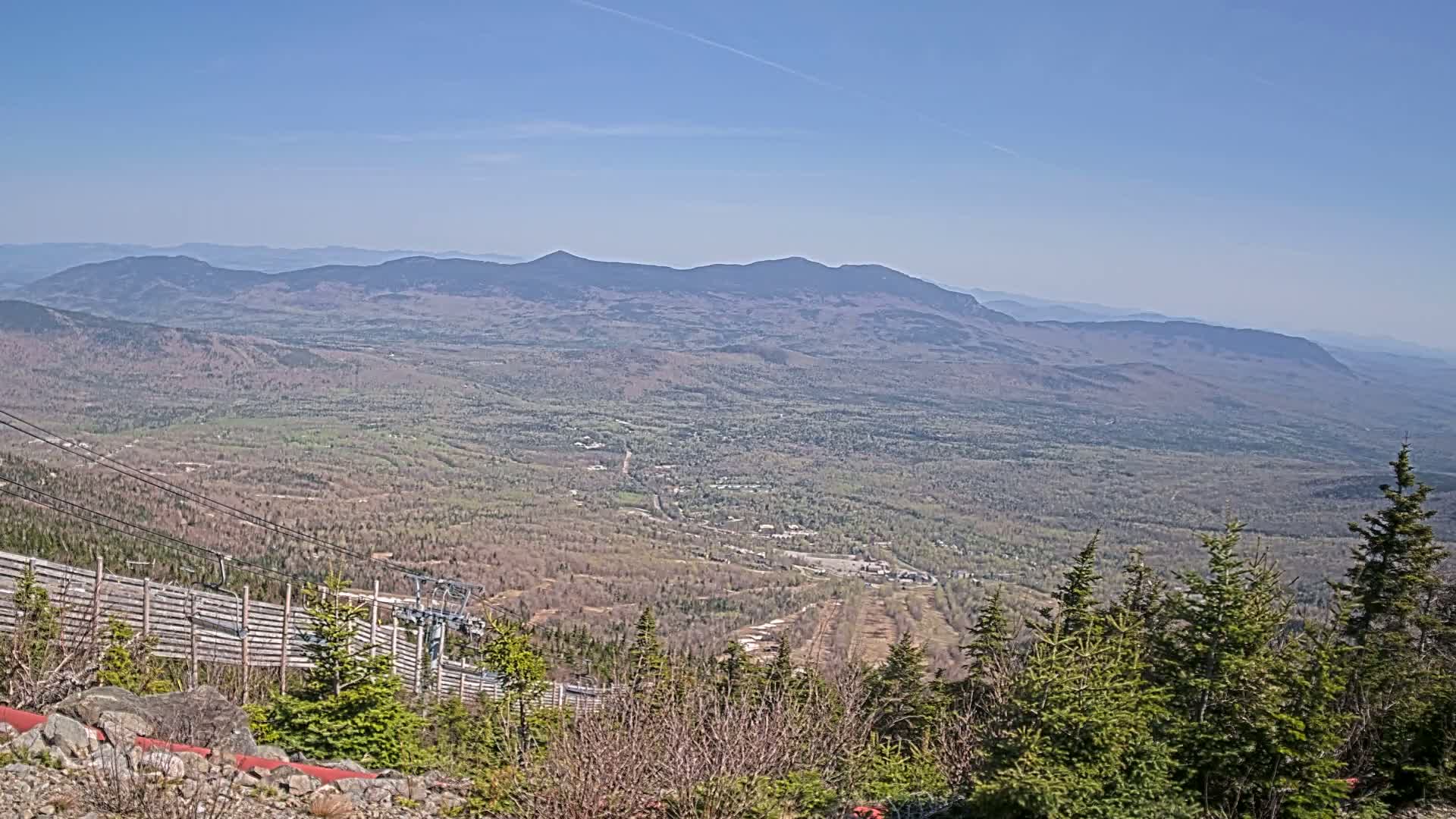 A vast, mountainous landscape under a clear blue sky stretches to the horizon, with a ski lift and wooden fence visible in the foreground.