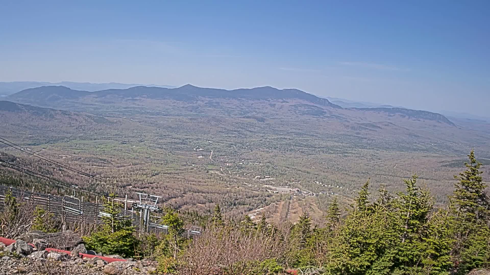 A high-angle, long shot on a clear day shows a vast expanse of forest and distant mountains, with a ski lift and part of a ski resort visible in the foreground.