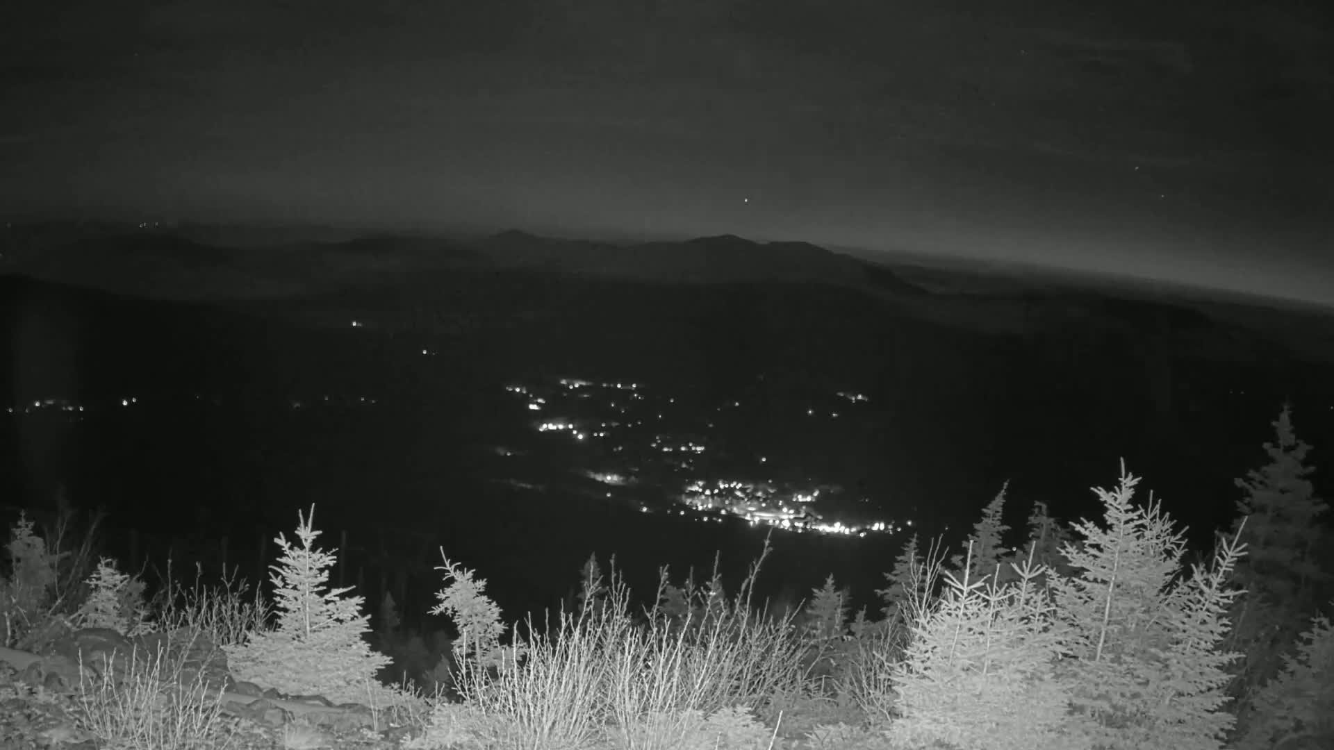 A nighttime view from a mountaintop shows a valley with scattered lights below, under a dark, clear sky.