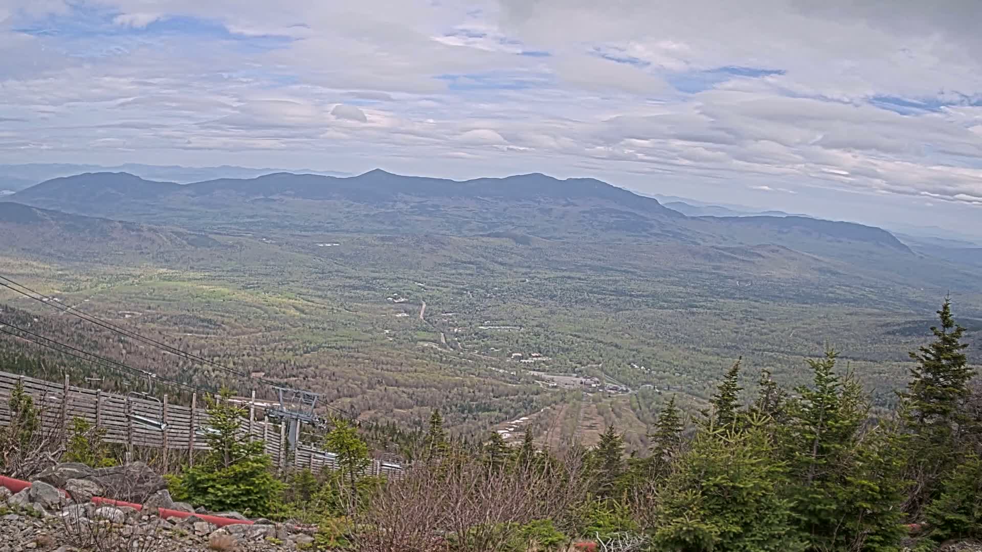 A sprawling vista of a mountainous valley blanketed in spring greenery under a partly cloudy sky is seen from a high vantage point, featuring a ski lift in the foreground.