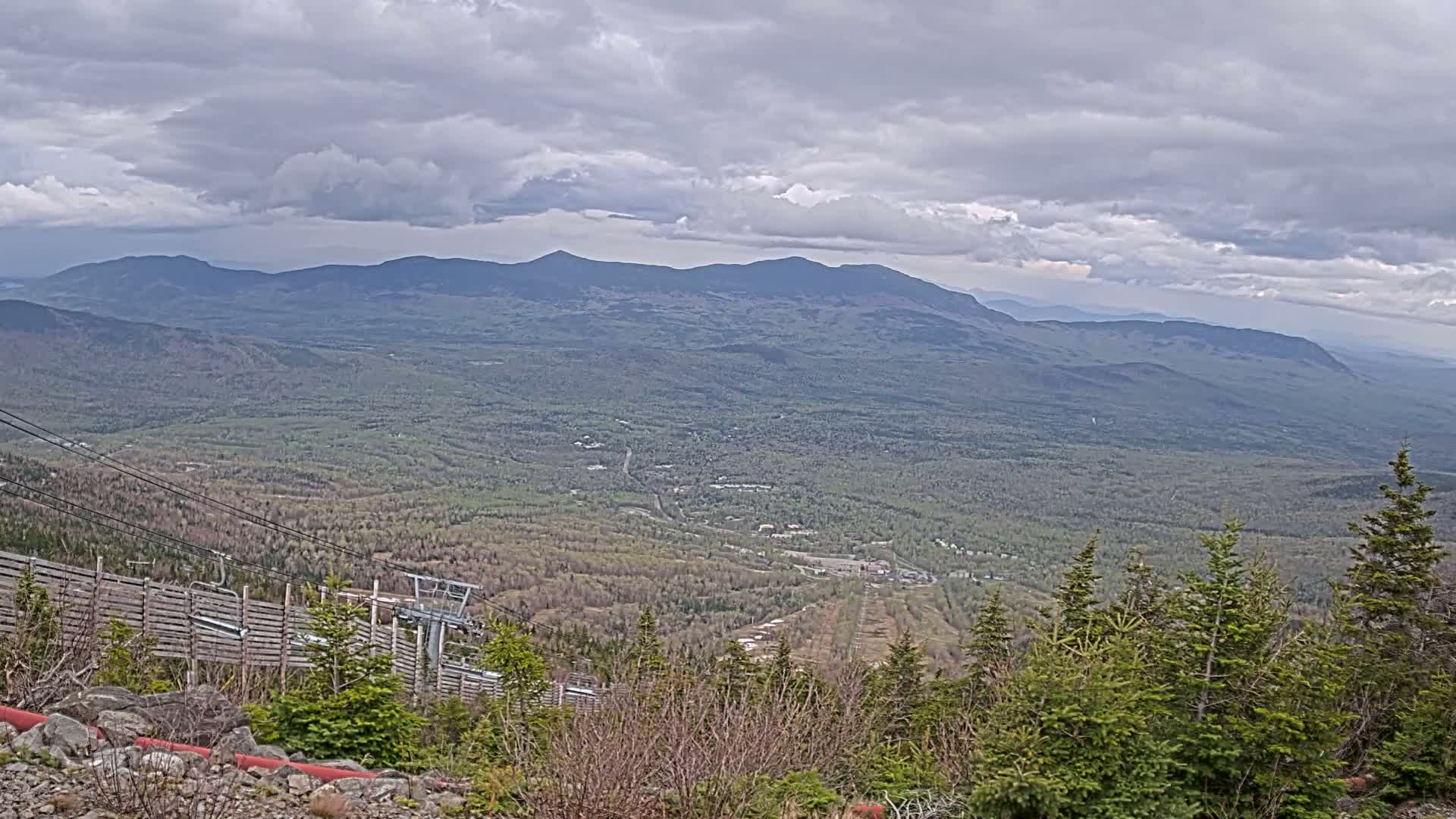 A mountaintop view under a cloudy sky reveals a vast expanse of forest and distant mountains, with a ski lift and wooden fence visible in the foreground.