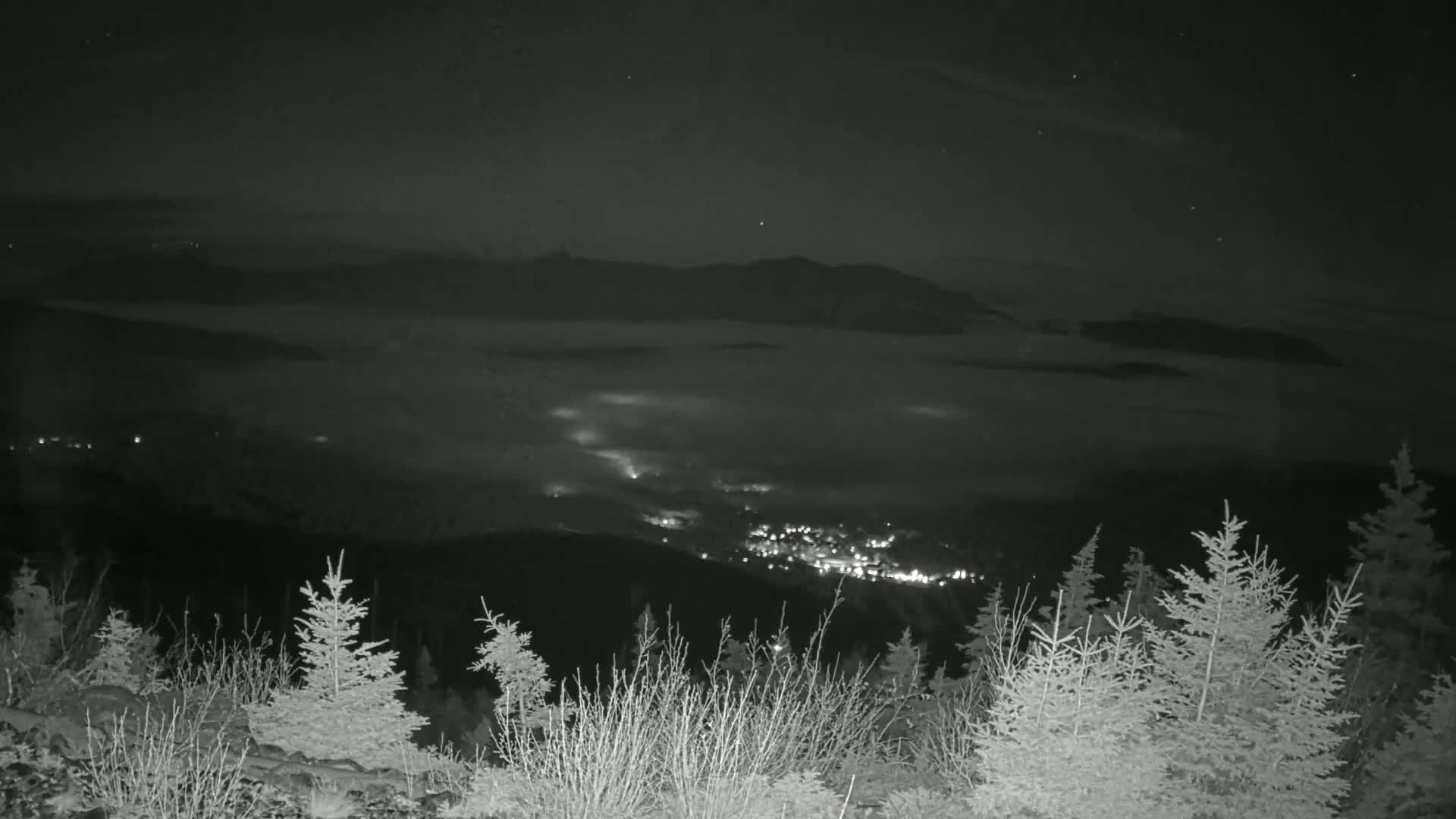 A nighttime view from a snowy mountaintop shows a valley below covered in a thick layer of fog, with a town's lights visible through it.