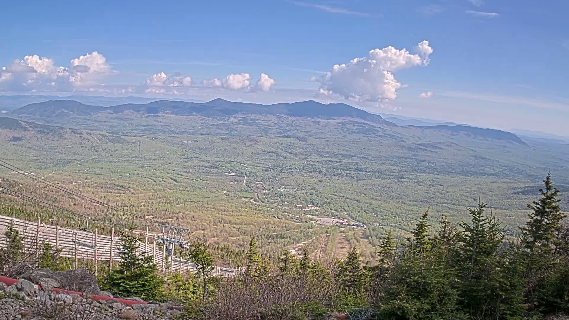 A vast, verdant mountain valley stretches to distant, hazy mountains under a partly cloudy blue sky.