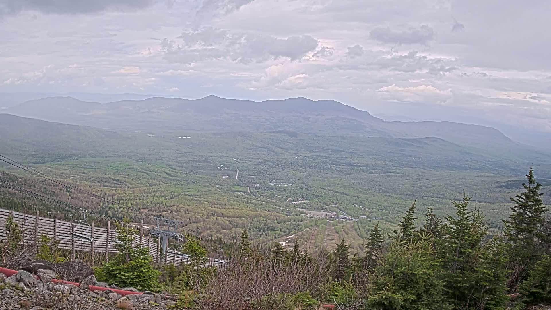 A mountainous landscape under a cloudy sky, featuring a ski lift and a valley covered in lush green vegetation.