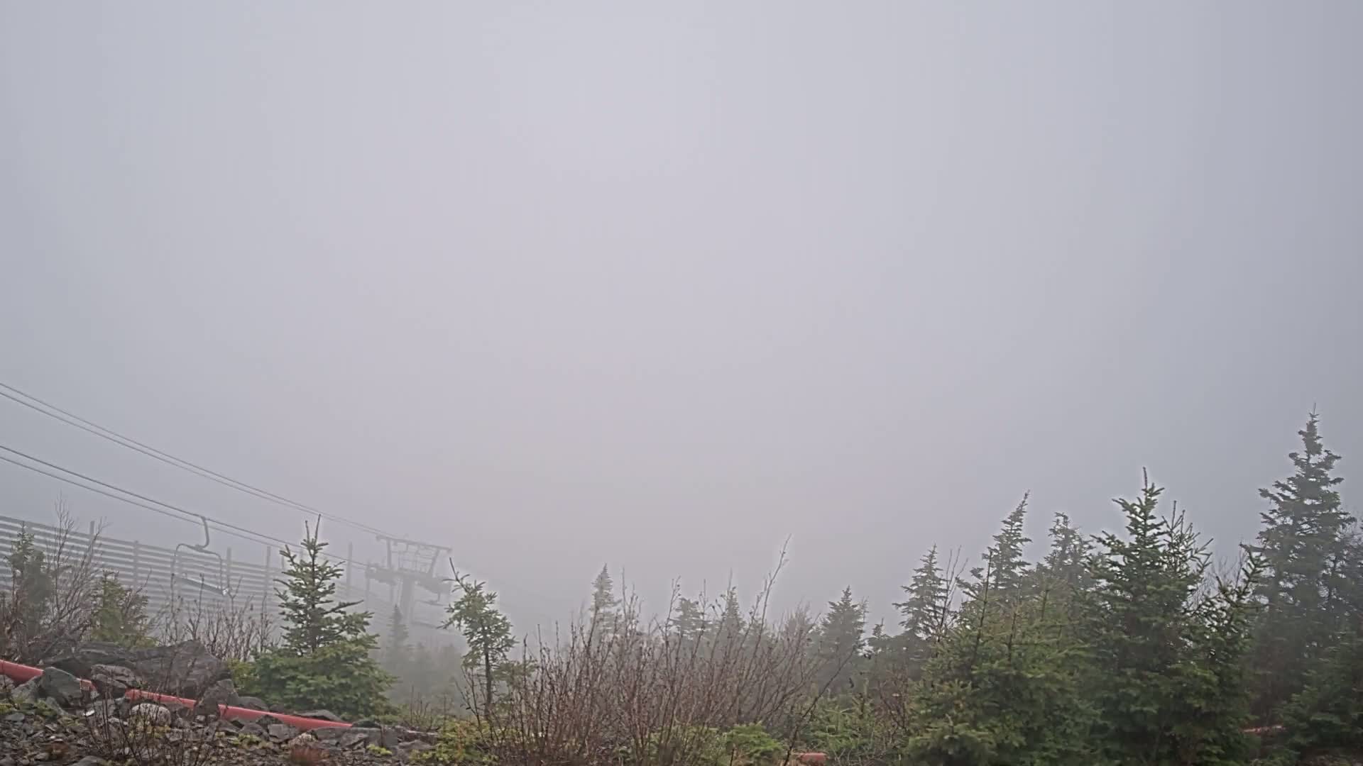 A thick fog obscures a mountaintop view of evergreen trees, a ski lift, and a wooden fence.