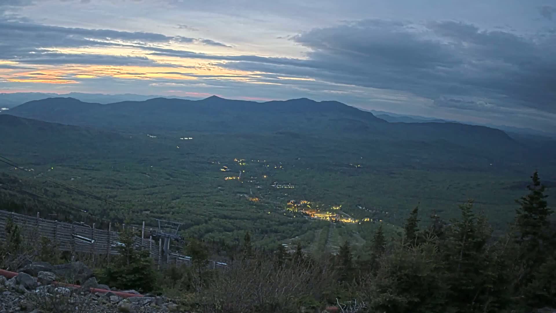 A mountaintop view at twilight reveals a valley with a town illuminated below, layered mountains in the distance, and a ski lift partially visible in the foreground under a partly cloudy sky.